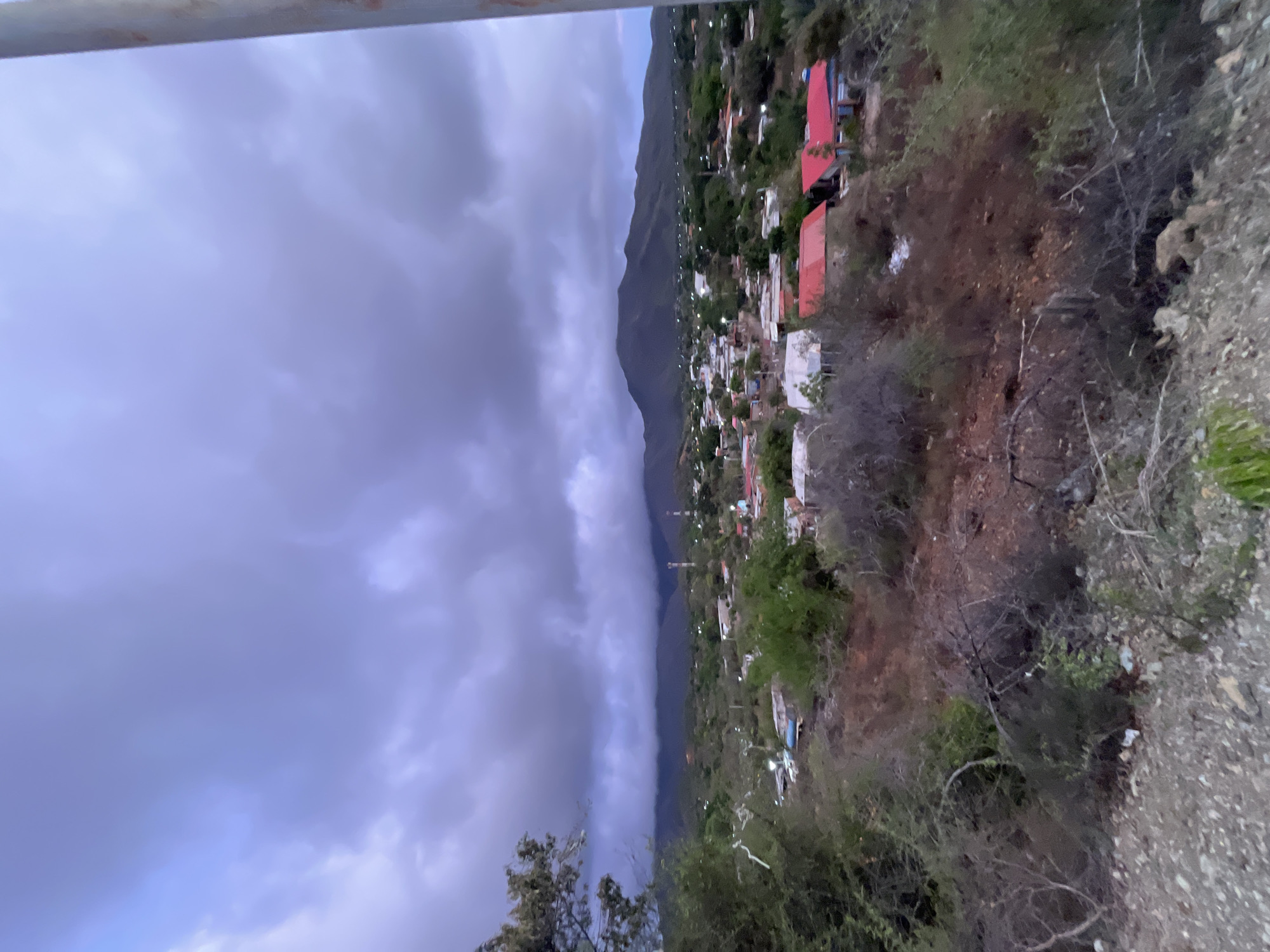 The image captures a panoramic view of a residential area in Santa Ana, Venezuela, under a dramatic, overcast sky. In the foreground, a rugged, dry hillside is covered in sparse vegetation and rocky terrain. Beyond this, a scattering of houses with red and blue roofs are visible, nestled amongst trees and other greenery. The middle ground features more houses and some taller structures, possibly communication towers, interspersed with dense foliage. In the distance, a range of dark, rounded mountains forms the backdrop, partially obscured by low-lying clouds. The overall impression is one of a remote, rural or semi-rural settlement during a potentially stormy or twilight period.
