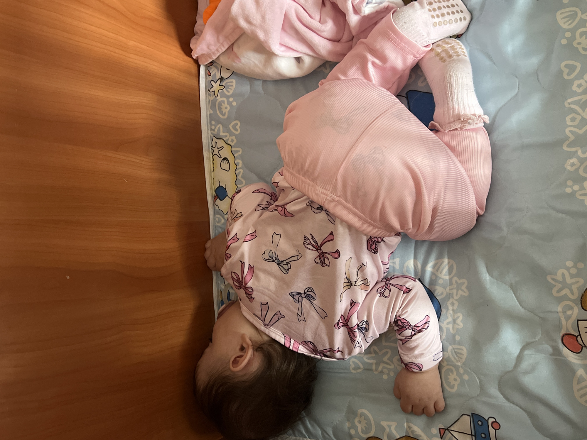 A baby is lying on a blue and white patterned baby blanket on a wooden surface. The baby is wearing a pink and white onesie with a bow pattern, pink pants, and white socks. The baby's head is turned towards the wooden surface, and the feet are raised towards the opposite side of the blanket. A pink blanket is partially visible at the top of the image. The scene is indoors. It is not possible to verify the location in the city of Elena, Bulgaria, from the provided image.