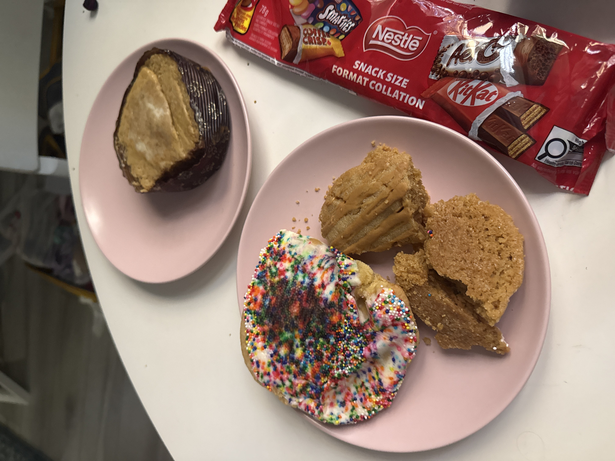 A top-down view of a white table is shown, with two pink plates and a red snack bag of candies. On the plate in the foreground, there is a circular cookie covered in white frosting and rainbow sprinkles, and two pieces of a broken cookie with a caramel-like drizzle. The plate in the background contains a single cupcake with a dark wrapper and a tan frosting. The red snack bag to the right contains a variety of Nestle products, including Kit Kat bars and Smarties. The lighting suggests an indoor setting, likely a home or cafe, with no discernible landmarks of Milan, Italy.