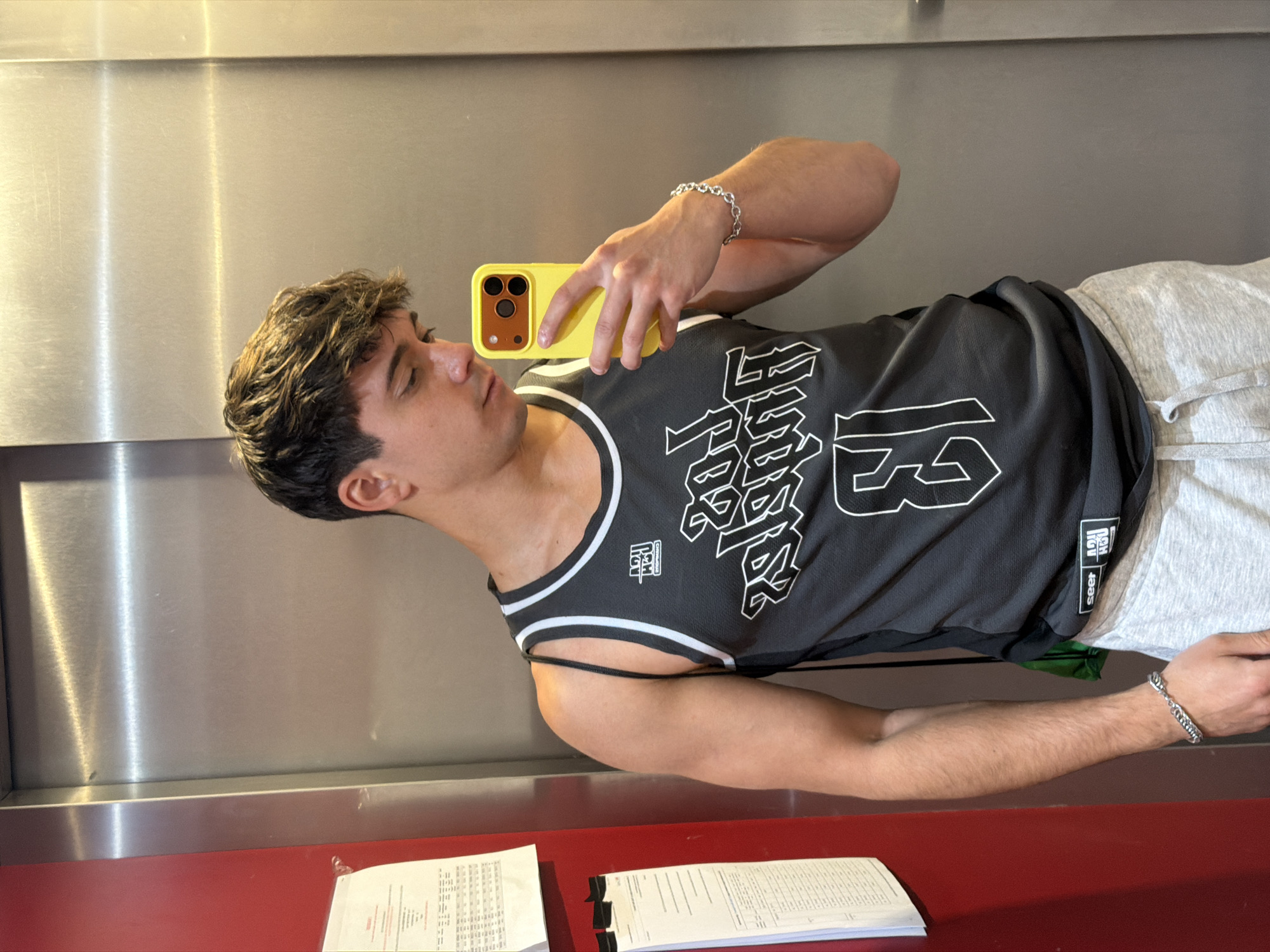 A young man takes a selfie in a reflective surface, likely in an elevator or a modern interior with brushed stainless steel walls, situated in Sofia, Bulgaria. He is positioned sideways in the image, with his head tilted slightly to the right. He wears a black athletic tank top or jersey featuring prominent white stylized Asian characters and the large number "13" on the front. A small tag on the side of his shirt displays the word "seer" and a logo that appears to read "HG RCH SPORTSWEAR". A silver chain bracelet is visible on his left wrist. He holds a smartphone with a vibrant yellow case and a triple camera system, directing his gaze towards its screen to compose the selfie. Below him, a red countertop or ledge holds several white documents with small, illegible text. The background consists of reflective brushed stainless steel panels, suggesting an indoor setting with artificial lighting. The man's expression is neutral and focused on taking the picture.