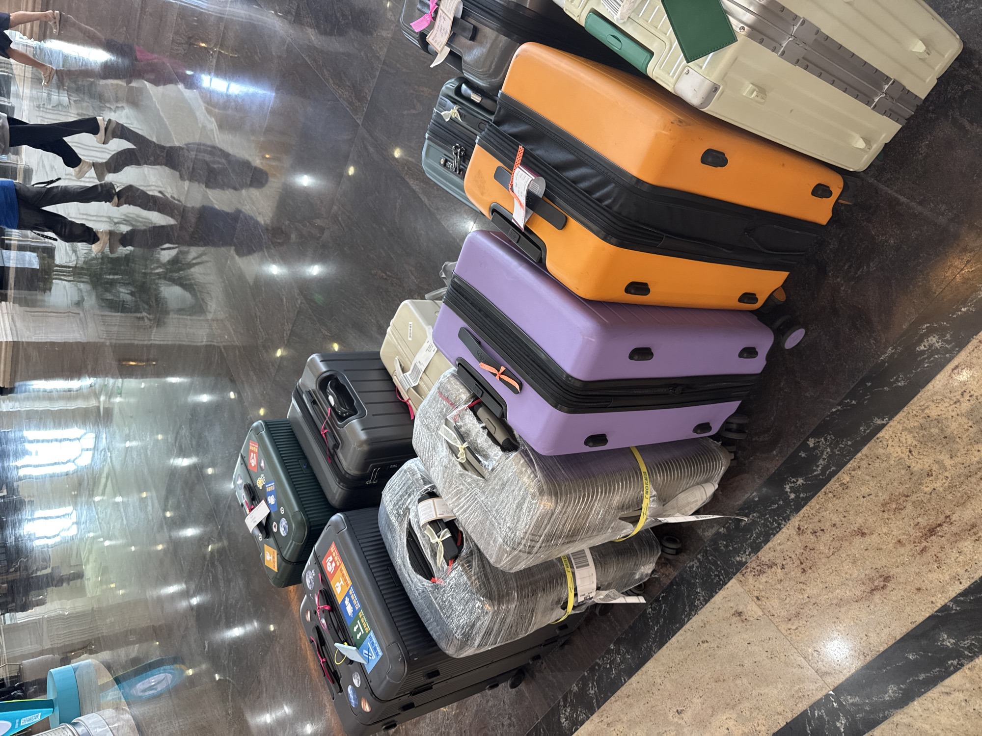 A stack of approximately ten pieces of luggage of varying colors, sizes, and conditions are piled on a shiny floor that reflects light and images of people and objects. The luggage includes orange, purple, silver, green, tan, and black suitcases. Some are wrapped in plastic, while others are adorned with stickers and tags. The floor appears to be tile or polished stone with a pattern, potentially inside a building, possibly in Nha Trang Ward, Vietnam. Reflected images suggest people moving in the area.