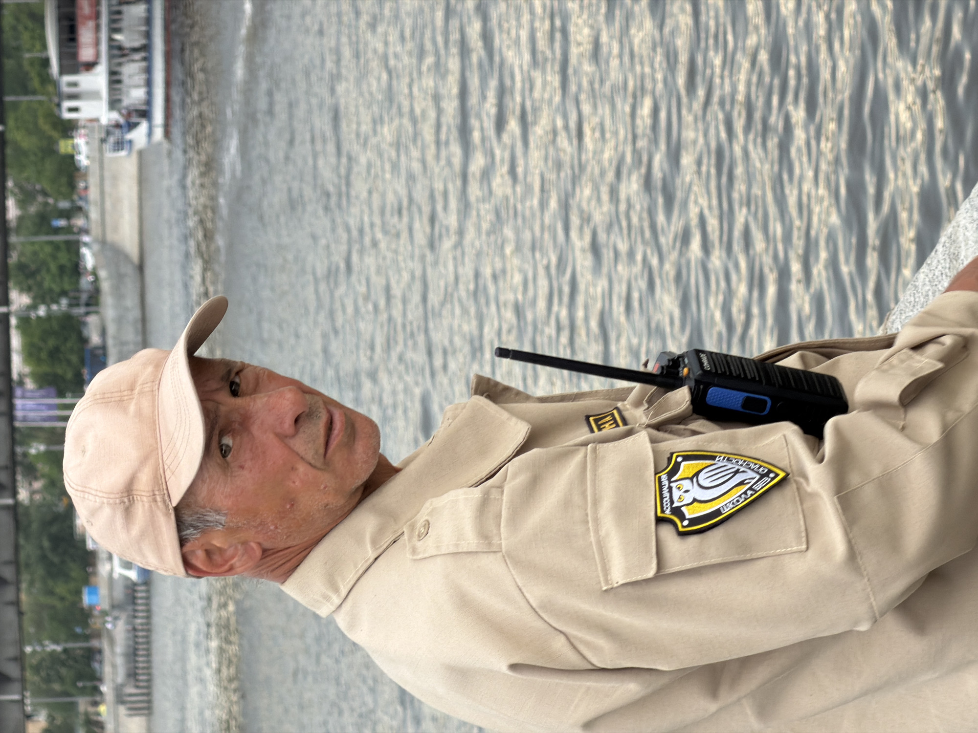 A man wearing a light-colored cap and uniform with a patch that appears to be the "Accolada Security School" logo is standing near a river. He has a black walkie-talkie clipped to his uniform. The background shows a river with sunlight reflecting on the water. On the far side of the river, there is a embankment. The ships are moored there. The embankment is covered with grass and trees. Based on the ships, the architecture, and the uniform patch, this scene could be in Moscow, Russia.
