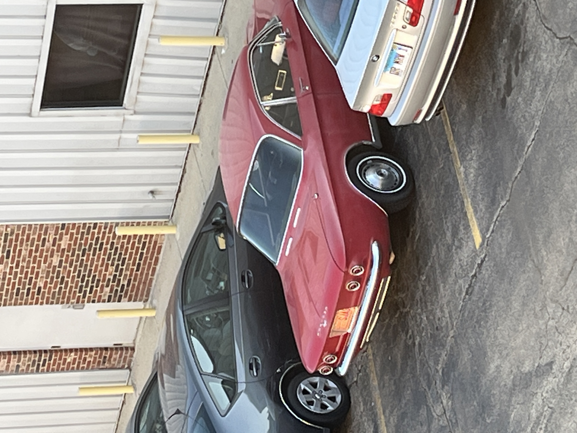 The image captures a parking scene in Woodridge, Illinois, United States. A red vintage car is parked next to a modern gray sedan and a tan BMW. The vehicles are in an exterior setting of a parking lot with yellow line markers on a gray concrete surface. The background features a building exterior with alternating sections of red brick and white metal siding, including a window. Yellow bollards are spaced intermittently along the building's base. No people are visible in the photograph.