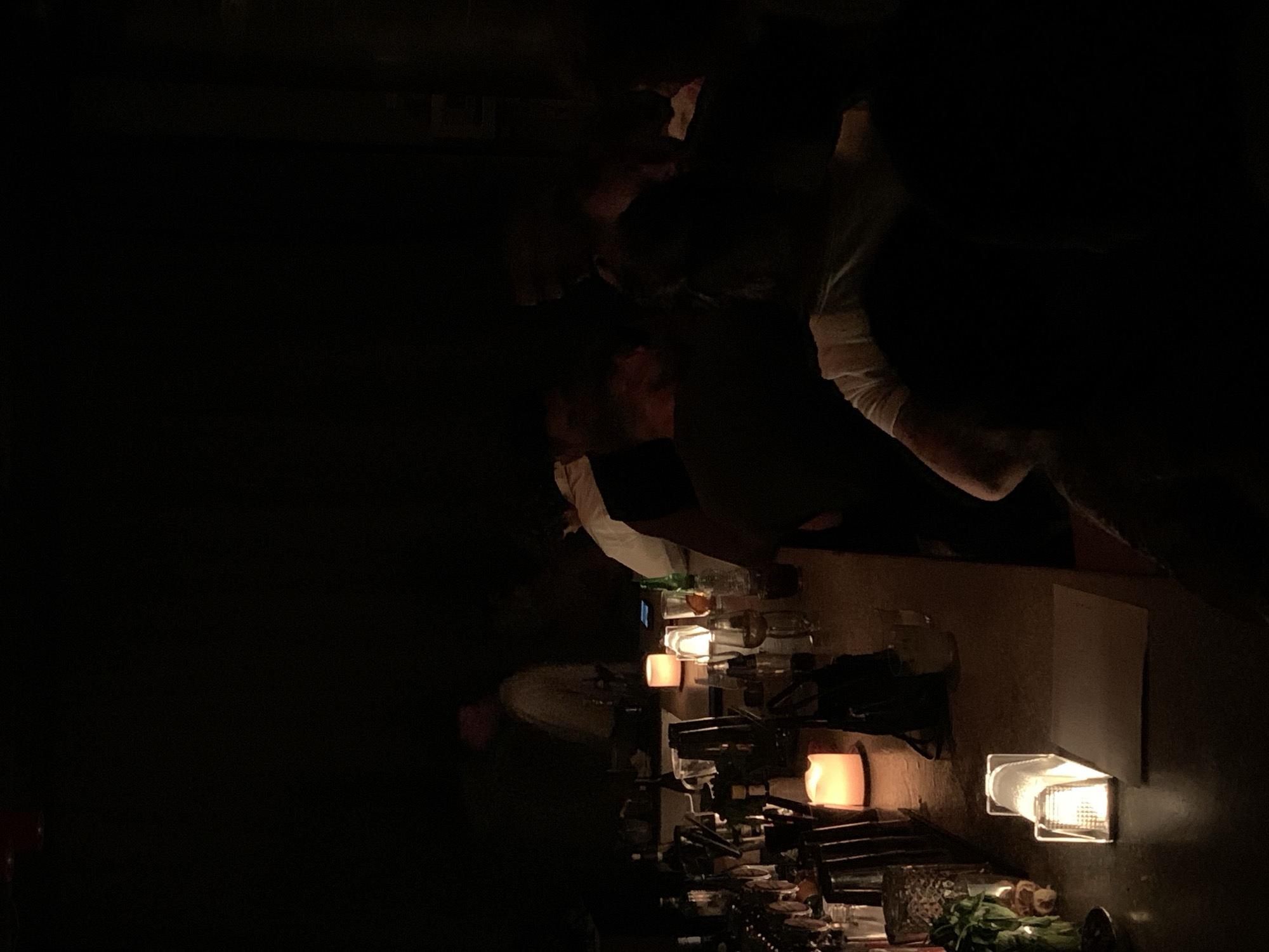 Here's a description of the image:
**Setting:** A dimly lit bar environment, possibly in Tel Aviv, Israel. Evidence suggests this is a bar due to the presence of glassware, bar equipment, and people interacting near the counter.
**People:** The image captures several individuals, though they are mostly obscured by the low lighting. The focus seems to be on the interaction at the bar. One person with a white sleeve is more prominently visible on the right side.
**Objects:**
* Various glassware and bottles are arranged along the bar top.
* Bar equipment is visible.
* Several light sources, including wall-mounted lights and what appear to be candles, provide illumination.
* A piece of paper or menu is mounted on the wall.
* There appears to be greenery, possibly herbs or garnishes, on the bar.
**Lighting:** The scene is heavily reliant on artificial lighting, which creates a warm, intimate atmosphere. The light sources are limited, leading to stark contrasts and deep shadows.
**Actions/Interactions:** People are interacting at the bar, though the specifics of their activities are difficult to discern due to the low light and obscured views.
The setting, with the bar and dim lighting, suggests a social environment, likely at night.