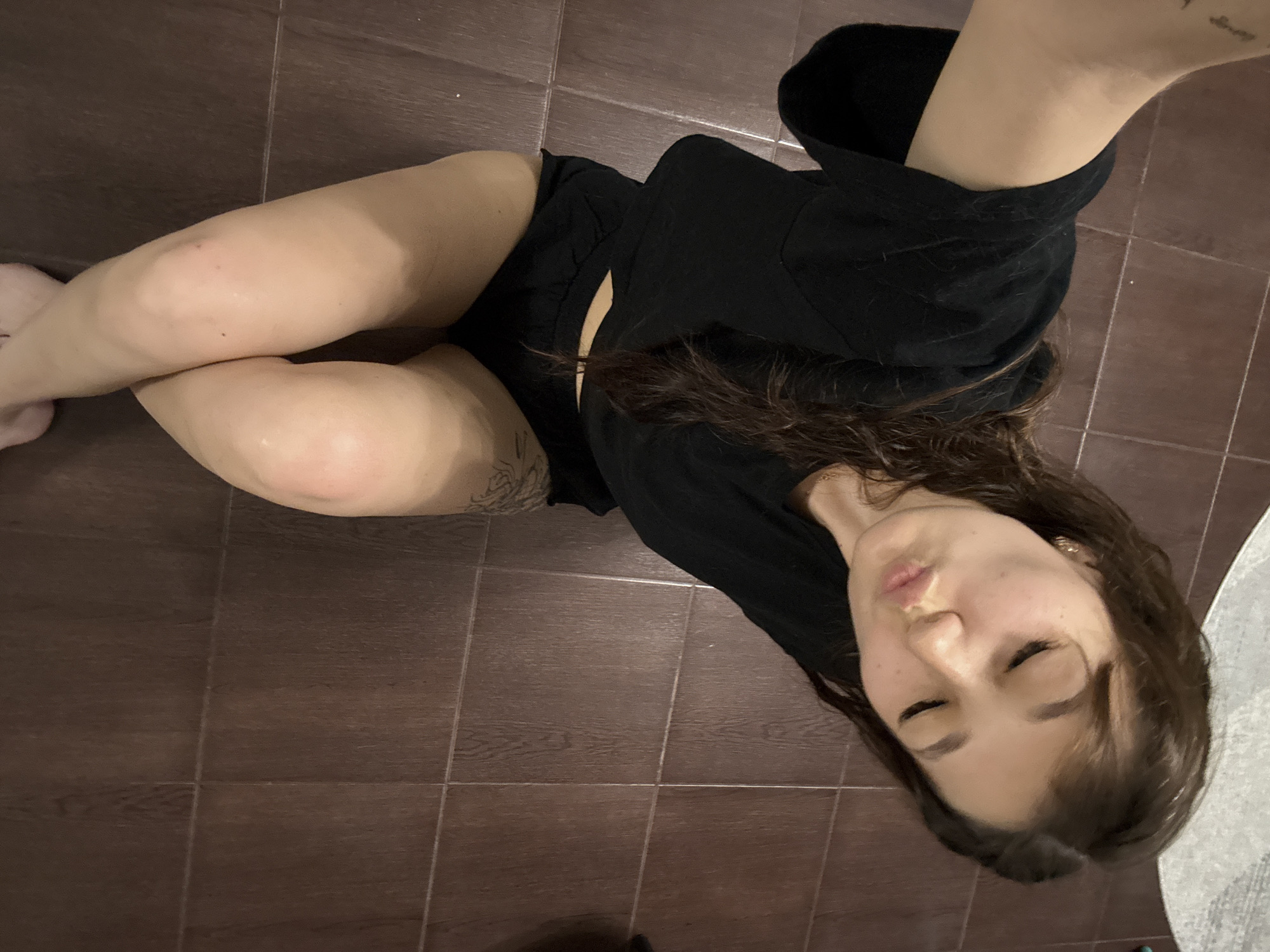 A young woman is lying on a tiled floor, posing for a selfie. She is wearing a black t-shirt and black shorts. Her eyes are closed and her lips are pursed in a kiss. A tattoo is visible on her left thigh. The floor is made of dark brown tiles. The image appears to be taken indoors. There is no information in the image to suggest a specific location in Novokuznetsk, Russia.