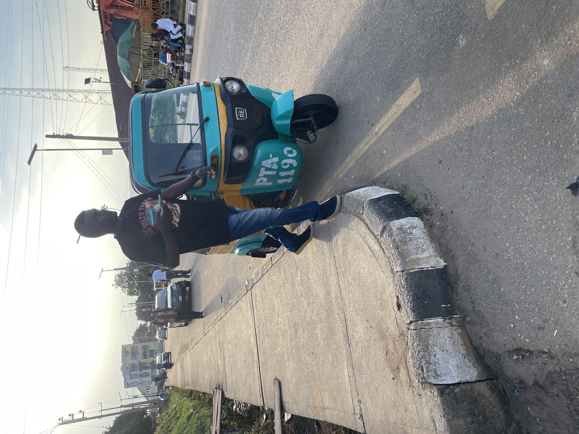 The image depicts a street scene in Jos, Nigeria, during what appears to be the late afternoon or early evening. A man, dressed in a black t-shirt and blue jeans, walks along a concrete sidewalk adjacent to a paved road. He is holding a small, rectangular object in his right hand and looking towards the road. To his right, a teal and yellow tricycle, commonly known as a "keke," is parked on the road. The tricycle has "PTA-1190" displayed on its side. Further down the road, several other vehicles, including cars and motorcycles, are visible. In the background, buildings, utility poles, and a large communication tower can be seen against a bright, overcast sky. A green umbrella shades a small seating area where several people are gathered. The sidewalk has a worn curb with alternating black and white stripes. The overall environment suggests an urban setting with ongoing traffic and pedestrian activity.