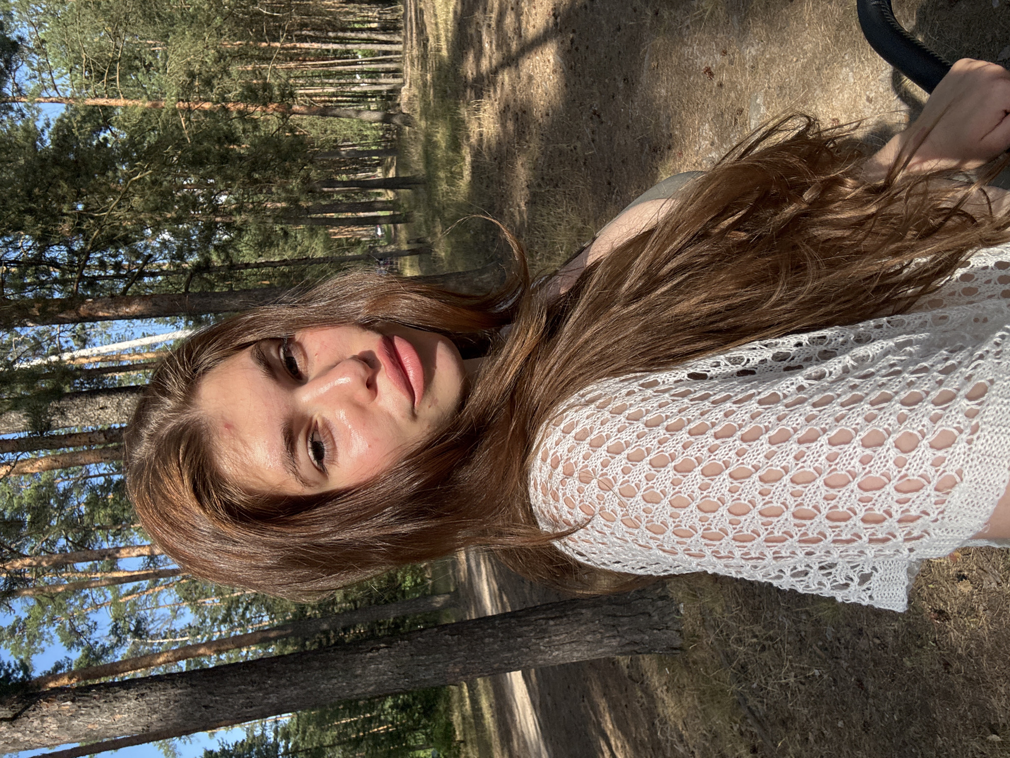 This media file shows a close-up portrait of a young person with long, light brown hair, lying down outdoors. The subject is facing the camera, with their head tilted to the side. Their eyes are open, and they appear to be looking directly at the viewer. Their lips are slightly parted, and they are wearing light pink lipstick. They are wearing a white, crocheted top that reveals glimpses of their skin through the open-knit pattern. The background is a forest, with tall pine trees and dry, grassy ground. Sunlight filters through the trees, casting dappled shadows. The overall impression is one of a relaxed and natural moment.
The subject's expression could be interpreted as calm or perhaps a little wistful. The lighting suggests it is daytime, likely a sunny afternoon, given the warmth of the light and the shadows. The presence of pine trees and dry, low vegetation is consistent with a forest environment in a temperate climate. The specific location of Kurovskoye, Russia, can be inferred, contributing to the context of a natural, wooded area. There is no visible text in the media file.