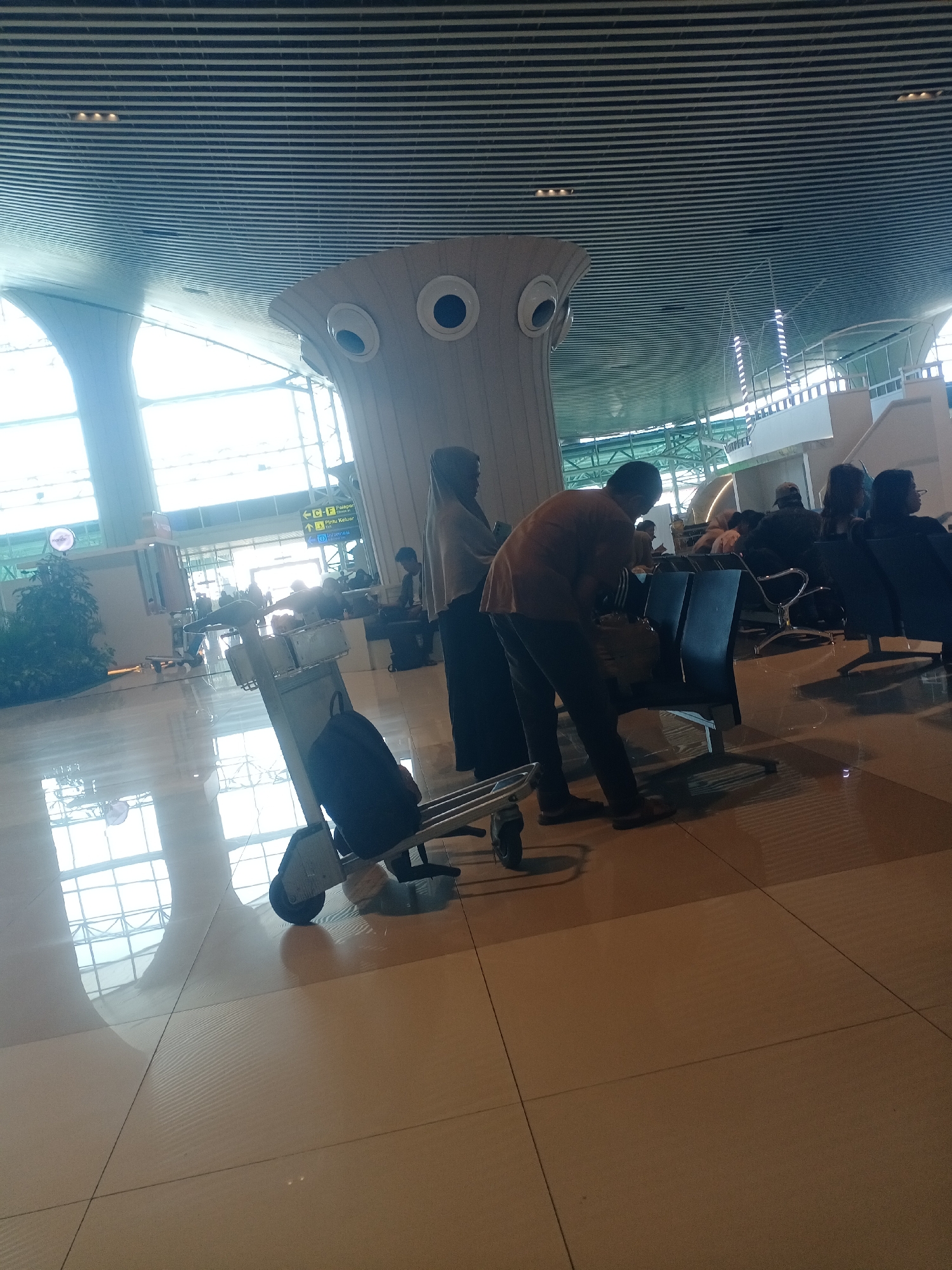 The image depicts an interior scene, likely an airport waiting area, given the presence of luggage carts, seating, and large windows. The architecture features a modern, curved ceiling with linear lighting and decorative elements resembling large eyes. The flooring is tiled and reflective, with clear reflections of the ceiling and windows. In the foreground, a luggage cart with a dark backpack on it is positioned prominently. Several individuals are visible in the midground and background, some seated and others standing. One man in an orange shirt and dark pants is leaning over a chair, while another person in a headscarf stands to the right. Signs with text, including "C-F" and "Pintu Keluar," are visible in the distance, suggesting a departure area. The overall lighting is bright, with natural light coming from the large windows. The scene is likely located within Baji Mangngai, Indonesia, inferred from the context of the prompt.