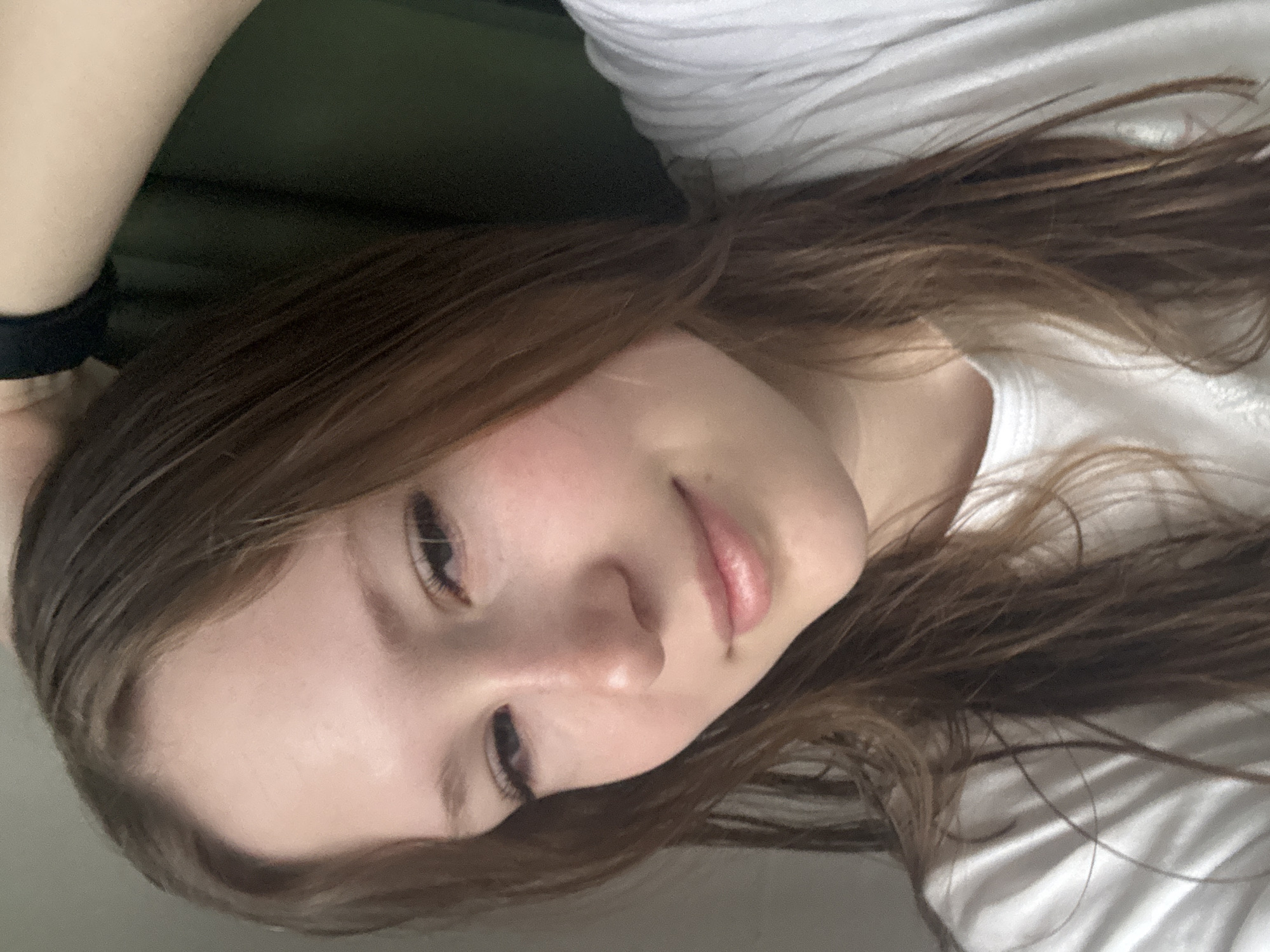 A young woman with light skin and brown hair is lying down. She is wearing a white t-shirt and a black band is visible on her left wrist. The background consists of a dark green surface. The specific location in Chaikovsky, Russia, is not discernable from the image.