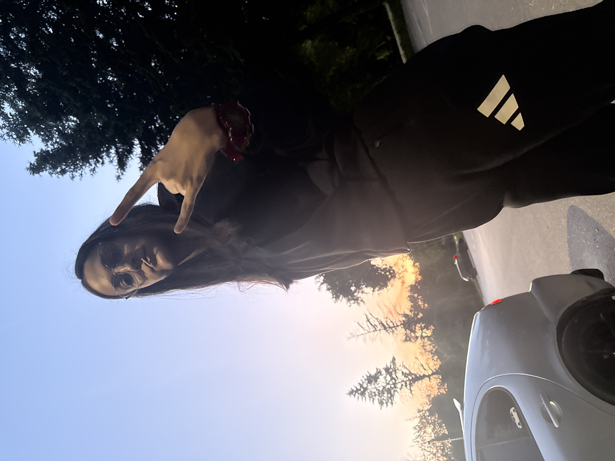 A person with long, dark hair and glasses is making a hand gesture with their left hand. They are wearing dark clothing with an Adidas logo on the leg. In the background, there are trees, a car, and the sky at sunset.
It is not possible to identify the location as Pleven, Bulgaria from the image.