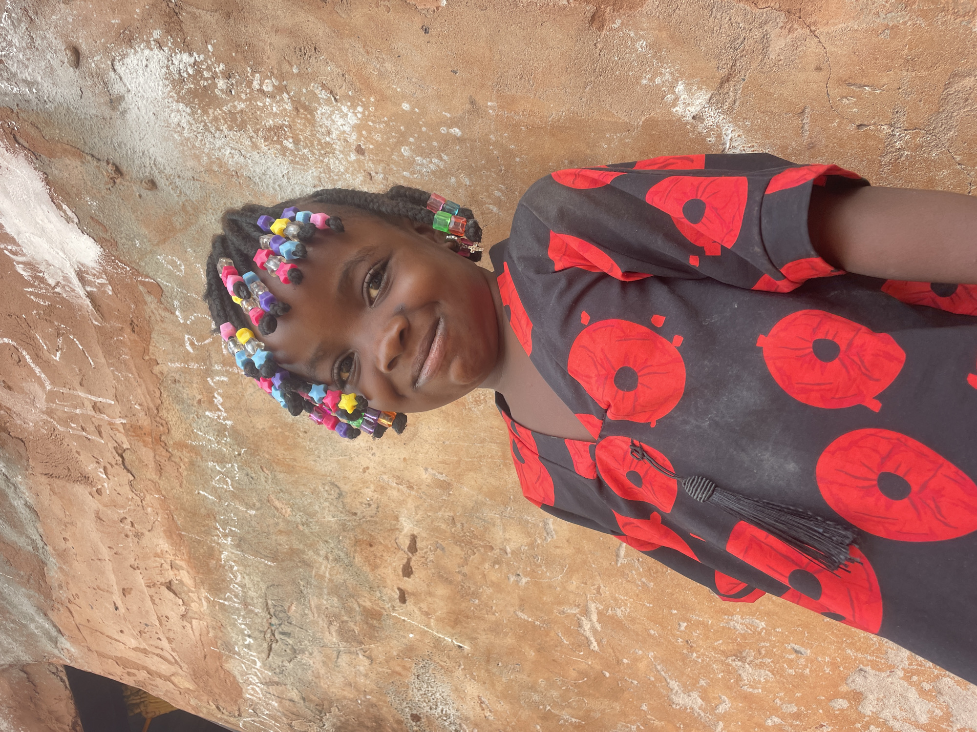 A young girl with braided hair adorned with colorful beads stands in front of a textured wall. She is wearing a dark-colored dress with a pattern of large red circular designs. The girl is smiling slightly, facing the camera. The wall behind her has a rough, earthy tone with patches of white and gray, suggestive of age and weathering. Based on the writing on the wall, this image was taken in Ogbe, Nigeria.