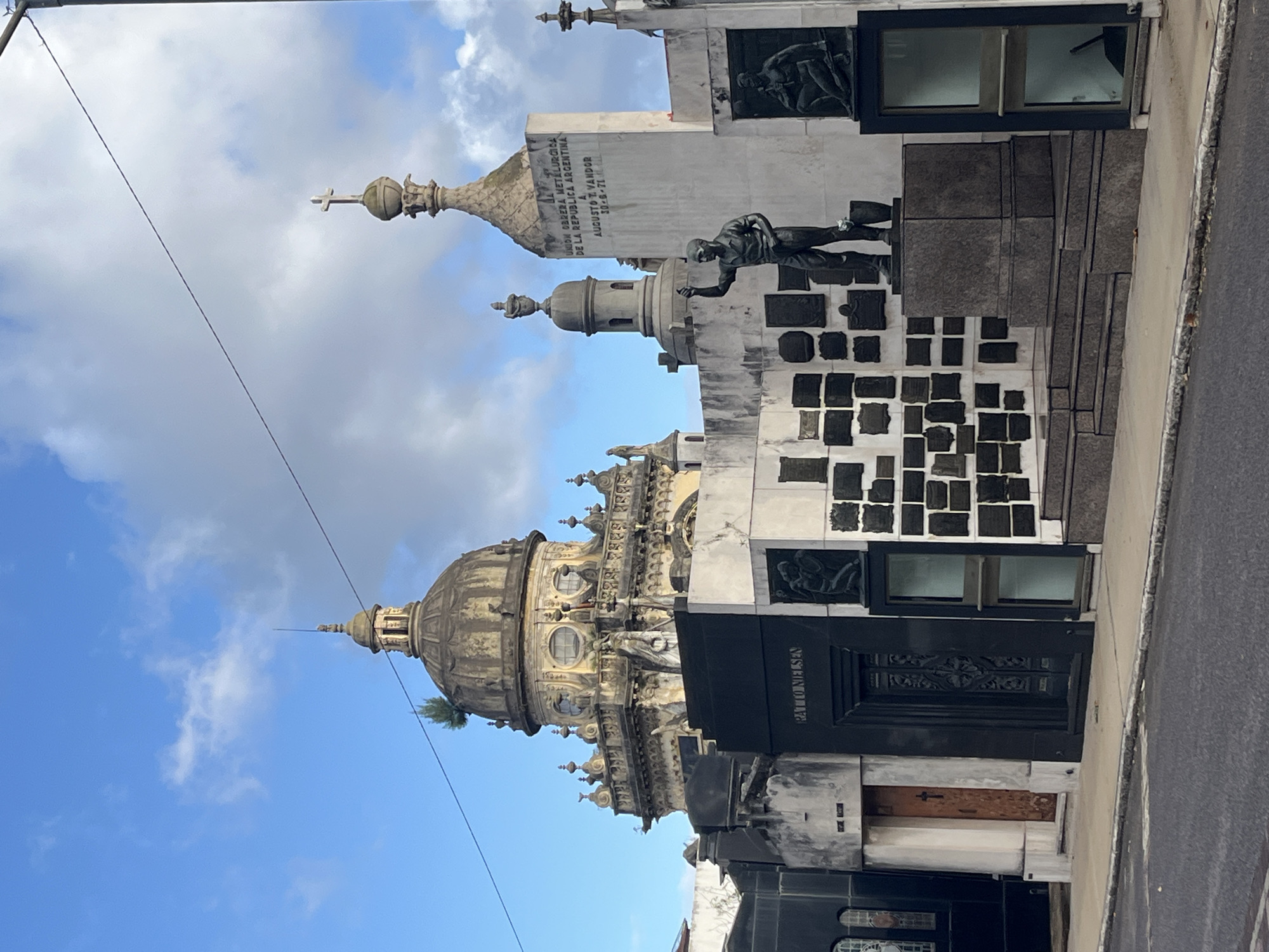 The image shows mausoleums at the Recoleta Cemetery in Buenos Aires, Argentina. The architecture features a domed structure, ornate decorations, statues, and smaller crypts or plaques affixed to the walls. The sky is blue with clouds. A portion of the street or walkway is visible on the right edge.