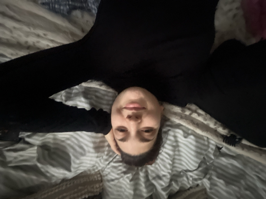 A person lies on a striped bed, facing the camera upside down. They wear a black long-sleeved shirt or bodysuit and stretch their arms out. Their head rests on their hand, with a slight smile on their face. A dark hair clip is visible on the bed near their right shoulder. The background is a blurred mix of bedding and possibly other items.
