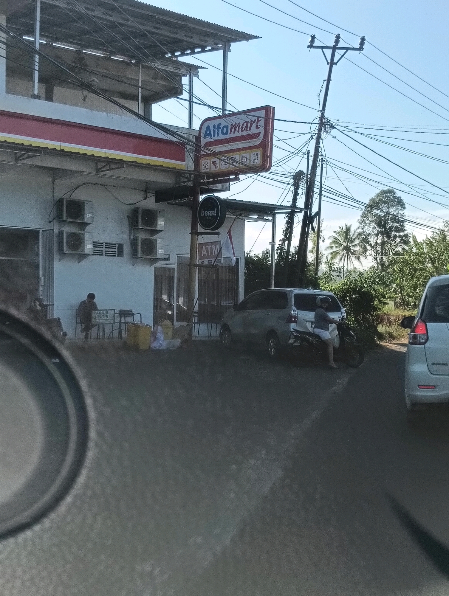The image captures a scene in Tomohon, Indonesia, featuring an Alfamart convenience store. In front of the store, several air conditioning units are mounted on the white wall. A person is seated on a chair outside the store, looking down at something on a table, with yellow and white plastic bags nearby. A sign that says "PARKIR GRATIS" (free parking) is visible on a stand.
To the right of the store, an ATM is advertised. Further down the street, a silver minivan is parked, and a person on a motorcycle is seated on it, facing away from the camera. A portion of another white car is visible on the far right. Overhead, a tangle of electrical wires crosses the sky, supported by utility poles. The background consists of lush green trees and vegetation, characteristic of the tropical environment. The sky is clear and blue. The foreground shows a blurred dark surface, likely the interior of a vehicle from which the photo was taken, with a circular dark object partially visible in the lower left corner.