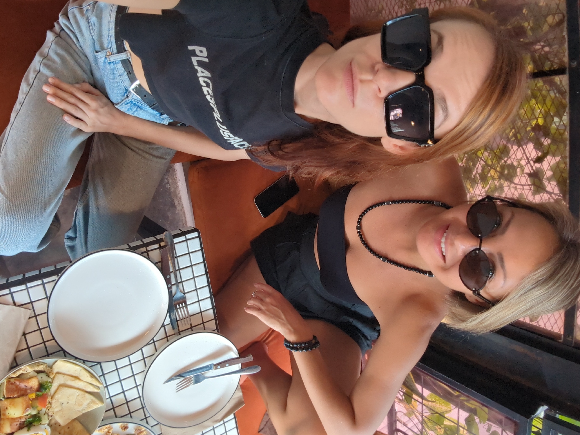 Two women are posing at a table covered in a black and white grid pattern, which is laden with plates and food. The woman in the foreground has blonde hair and wears a black top and sunglasses, along with a beaded necklace and bracelet. She is smiling. The woman in the background wears a black shirt, jeans, and large sunglasses. A smartphone rests on the orange bench between them. Based on information tags, the location is North Kuta, Indonesia.