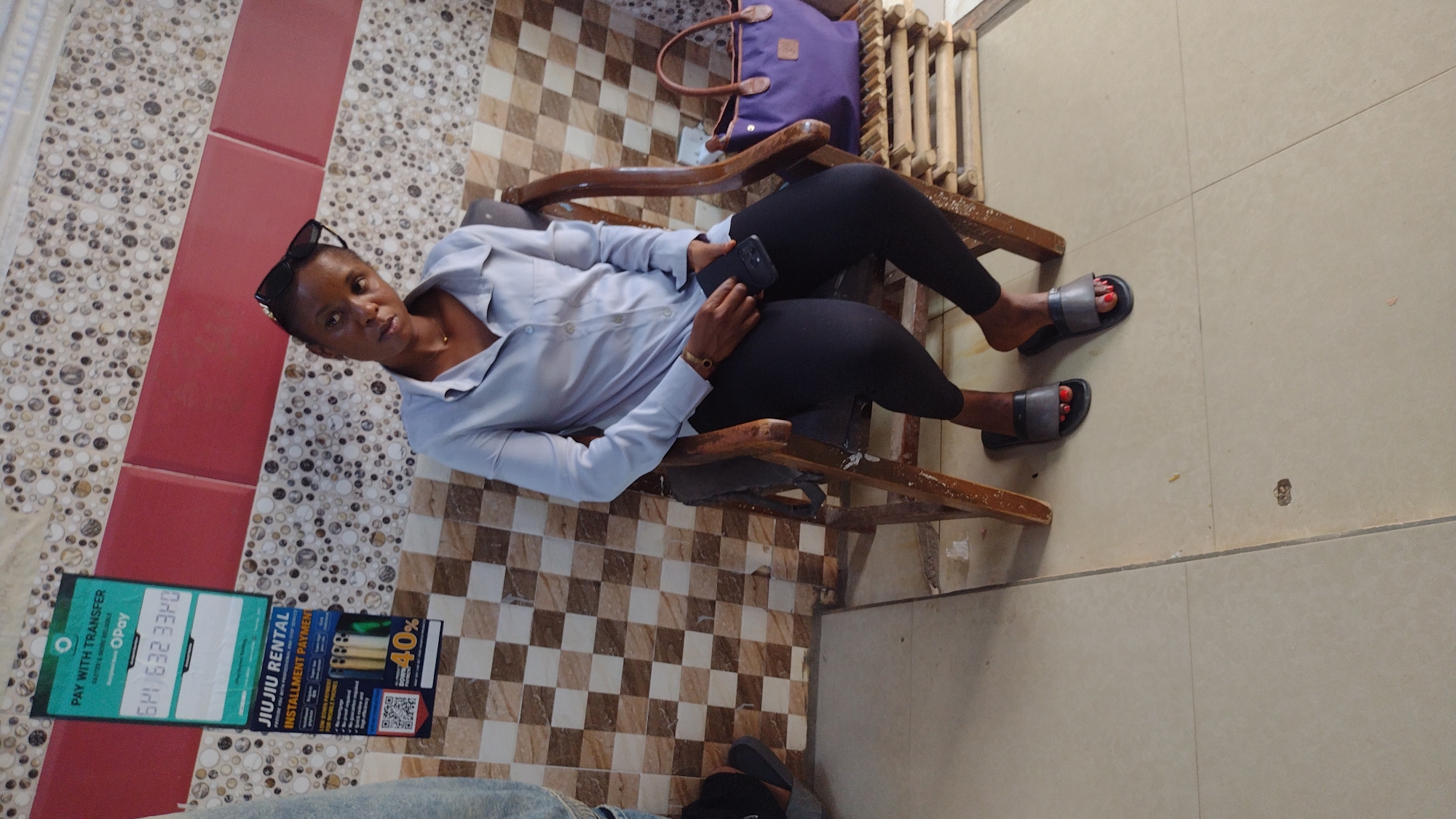 A woman sits in a wooden chair, wearing a light blue button-down shirt, black leggings, and dark gray slide sandals with red toenails. She is holding a dark-colored smartphone in her lap. Sunglasses are perched on her head. A purple bag is on the floor behind her chair. The floor is covered in a mixture of tile patterns, including speckled and checkered designs. A sign in the lower left corner advertises "JIUJIU RENTAL INSTALLMENT PAYMENT" and mentions "PAY WITH TRANSFER". The location, based on the sign's content and other visual elements, is likely Jos, Nigeria.