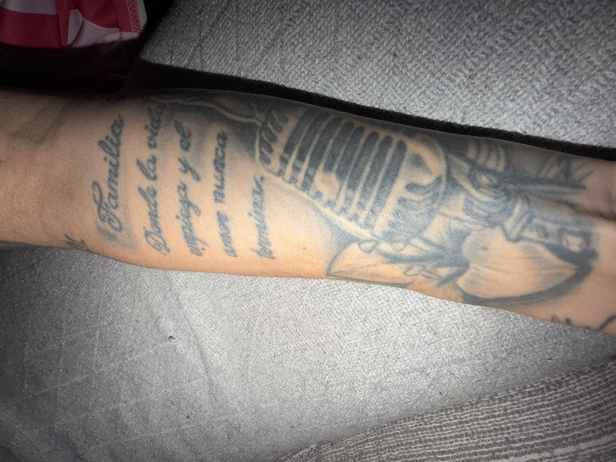 A forearm displays a tattoo with a microphone image accompanied by Spanish text that reads "Familia Donde la vida espiga y el amor nunca termina". The skin tone of the arm is light. The arm rests on a gray textured fabric.
