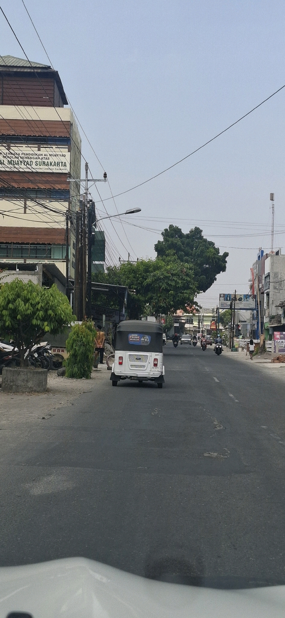 The image captures a street scene in Surakarta, Indonesia, with a white three-wheeled vehicle, commonly known as an "odong-odong," as the primary subject in the foreground, traveling away from the viewer. The vehicle has a black roof and a sticker on the back that reads "PAING MUSIM." The street is asphalted and appears to be a typical urban road with some potholes. On the left side of the road, there is a building with a sign that says "LEMBAGA PENDIDIKAN AL MUAYYAD SEKOLAH MENENGAH ATAS AL MUAYYAD SURAKARTA," indicating it is an educational institution. Several trees and a concrete planter with parked motorcycles are also visible along the left side. On the right side of the street, there are more buildings, including a telecommunications tower in the distance. Other vehicles, mostly motorcycles, are seen on the road, with some individuals present on the sidewalks. The sky is overcast, suggesting a neutral lighting condition.