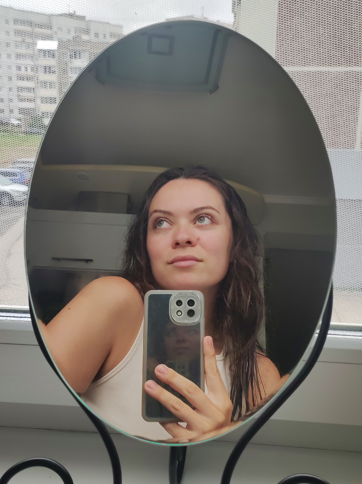 A woman takes a selfie in a round mirror. The reflection shows her holding a smartphone with a triple-lens camera. She has dark hair, fair skin, and is wearing a light-colored tank top. The background reflected in the mirror shows a room with white cabinets and a ceiling light. Outside the window, a building with many windows is visible, implying an urban setting. A mesh screen is visible on the window. Based on the architectural style of the building, the presence of the mesh screen, and the mention of Chaikovsky, Russia, the photo likely originates from an apartment building in that location.