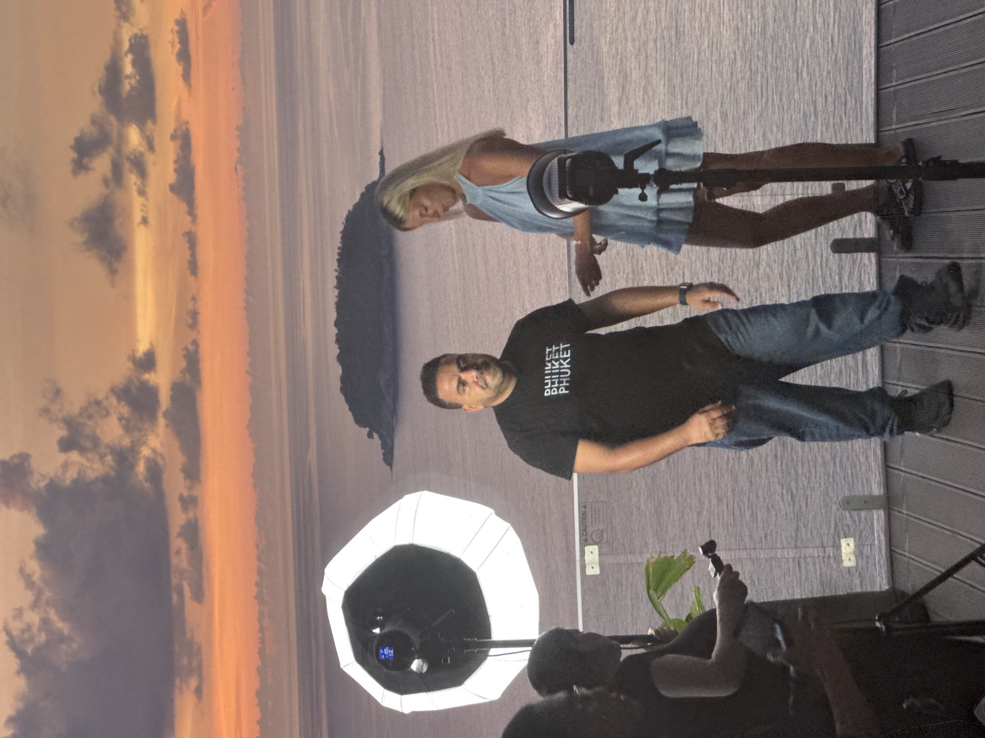 At sunset in Karon, Thailand, two people are being photographed. A man in a black t-shirt with the word "PHUKET" printed on it multiple times and blue jeans stands to the left. To his right, a woman in a light blue dress with ruffled hem stands. A large octagonal softbox light is positioned to the left of the man, and a smaller, cylindrical flash unit is placed to the right of the woman. They are on a wooden deck overlooking the ocean, with a small island visible in the distance. The sky is filled with dramatic clouds illuminated by the setting sun in shades of orange and pink.
