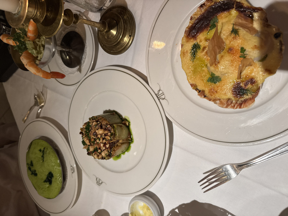A table setting displays four dishes: a shrimp cocktail in a brass container, a green purée, an artichoke heart topped with nuts, and a baked scallop in its shell. A silver fork lies beside the scallop dish. The setting includes a white tablecloth and ornate dishware with a logo.