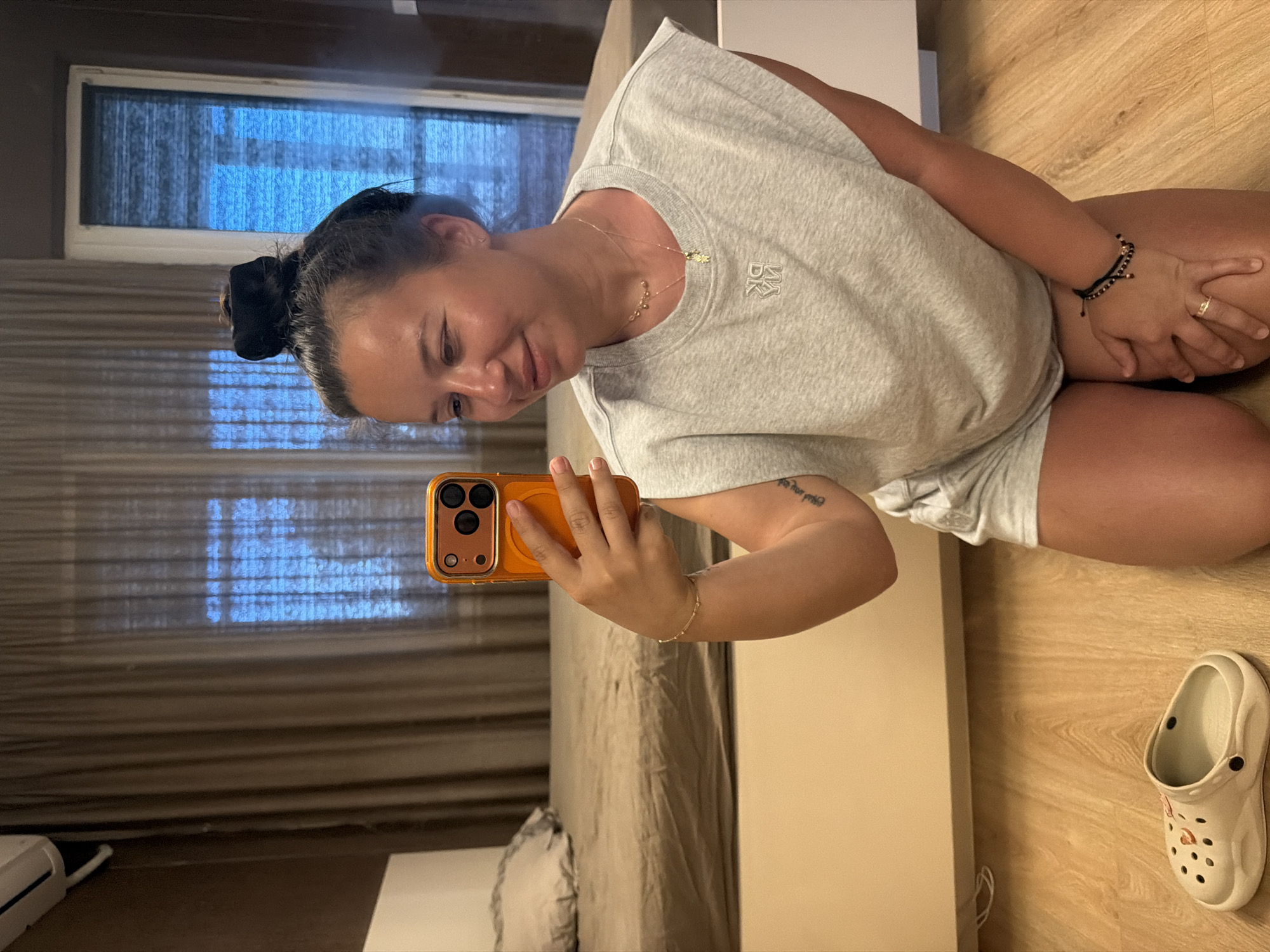 A woman is kneeling on a wooden floor, taking a selfie with her phone. She is wearing a grey t-shirt and shorts, with her hair pulled up in a bun. A pair of white Crocs are on the floor to her left. In the background, there is a bed with a beige comforter and a window with beige curtains. An air conditioner is mounted on the wall above the window. The scene appears to be an indoor domestic setting, likely a bedroom. This image was taken in Burgas, Bulgaria.