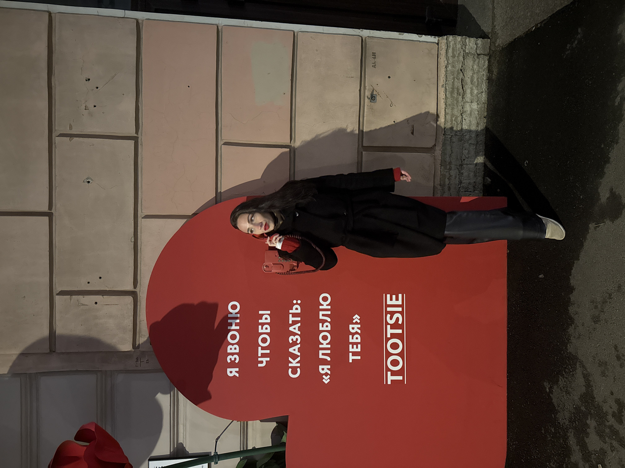 A sideways eye-level shot shows a woman in a black coat, black pants, and light-colored shoes lying on a large red telephone booth prop. The woman has dark hair and wears red lipstick. She is holding a red telephone receiver to her ear. The telephone booth has white Cyrillic text on it, and the English word "TOOTSIE" is printed in white. The text reads, "I am calling to say: I love you." The telephone booth is in front of a light pink, tiled building. The street is wet. There is Cyrillic text on the building. The location is most likely Saint Petersburg, Russia.