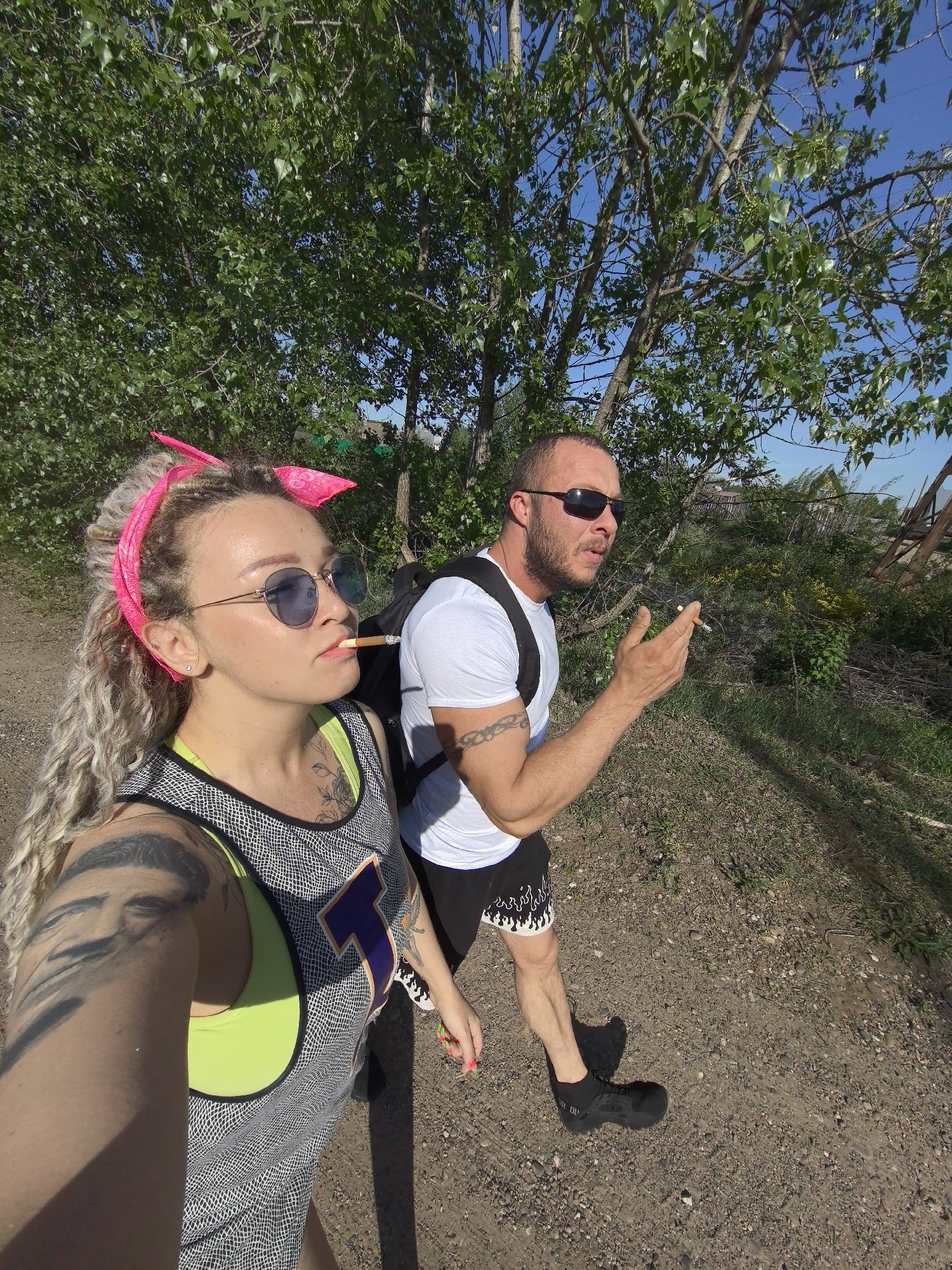 A couple is walking on a dirt path under trees. The woman on the left wears sunglasses, a pink headband, a patterned tank top with a neon green sports bra underneath, and has tattoos on her arm. She has a cigarette in her mouth. The man on the right wears sunglasses, a white t-shirt, black shorts with white flame patterns, and black shoes. He has a backpack and a tattoo on his arm. He is holding a cigarette in his hand and gesturing. The location is near Noviy, Russia, indicated by terrain and vegetation.