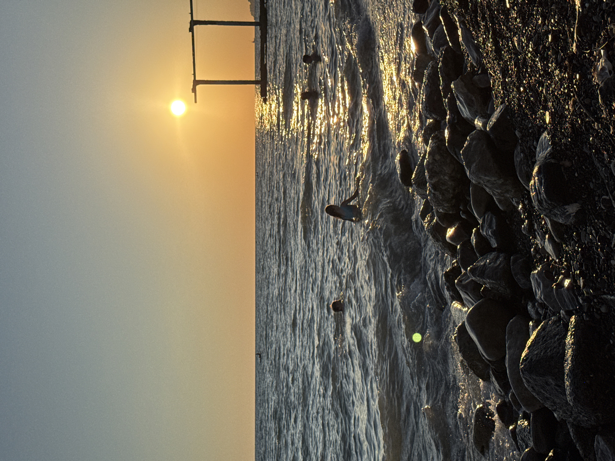 The image captures a serene sunset over a rocky beach, likely in Sochi, Russia. The sun, a bright orb, hangs low in the sky, casting a warm, golden glow across the scene. The sky transitions from a pale blue near the horizon to a soft, peachy orange as it ascends. The sun's rays glint off the gentle waves of the sea, creating a shimmering path of light.
On the beach, a collection of dark, irregularly shaped rocks forms a textured border to the water. Some of these rocks are wet, reflecting the sunlight. In the water, a single person is visible, appearing to be either wading or possibly bodyboarding, with their form silhouetted against the bright sea. The waves are small and appear calm, suggesting a tranquil atmosphere. The overall mood is one of peacefulness and natural beauty, enhanced by the warm light of the setting sun. The time of day is clearly sunset, with the soft light and long shadows indicative of the golden hour. There are no visible text elements in the image.