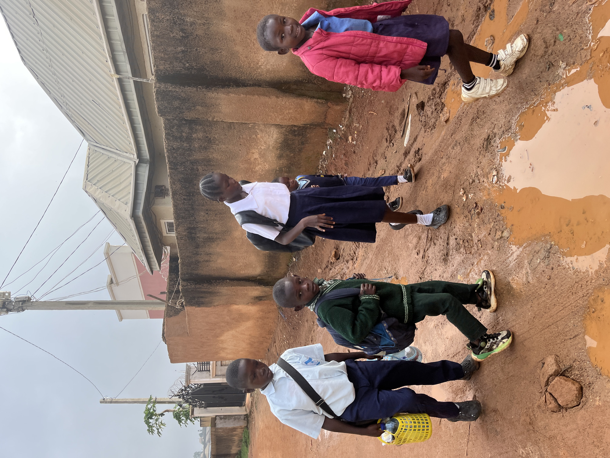 Five children, dressed in school uniforms, walk along a muddy, dirt road in Jos, Nigeria. The children appear to be walking away from the camera. The road is wet and has several large puddles of water. In the background, there is a low, brown wall, a gated entrance, and buildings. A utility pole with wires runs along the side of the road. The sky is overcast. The children are carrying backpacks and one child is carrying a yellow basket with bottles.