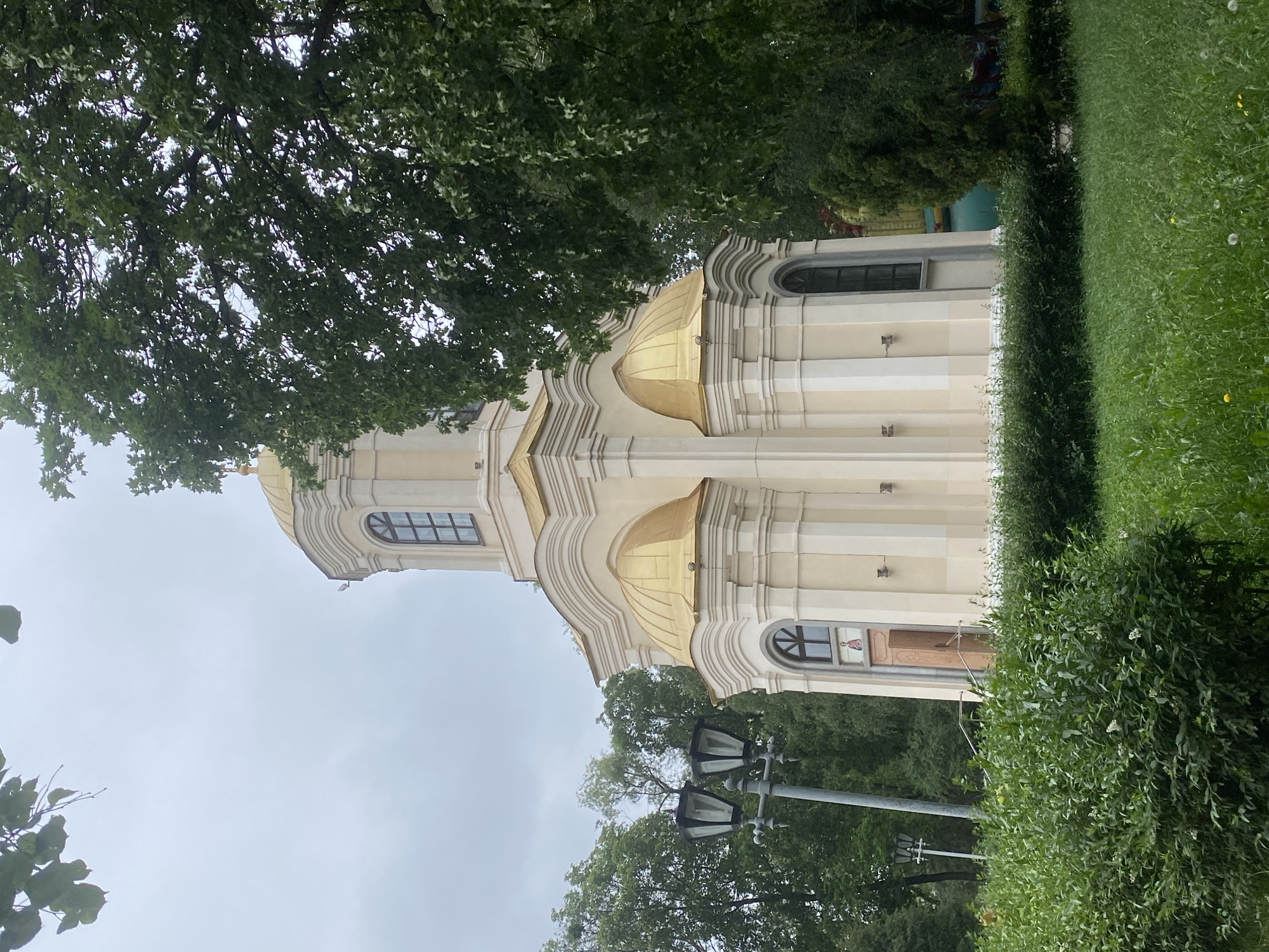 The image depicts a light-colored, domed church with golden cupolas, set against a backdrop of lush green trees and an overcast sky. The church features arched windows and an entrance with an open wooden door, above which an icon is visible. A lamppost with two lights stands to the left of the church, partially obscured by dense green bushes in the foreground. The church is located in Poltava, Ukraine. There are no people or animals visible in the image. The environment appears to be a park or a churchyard.