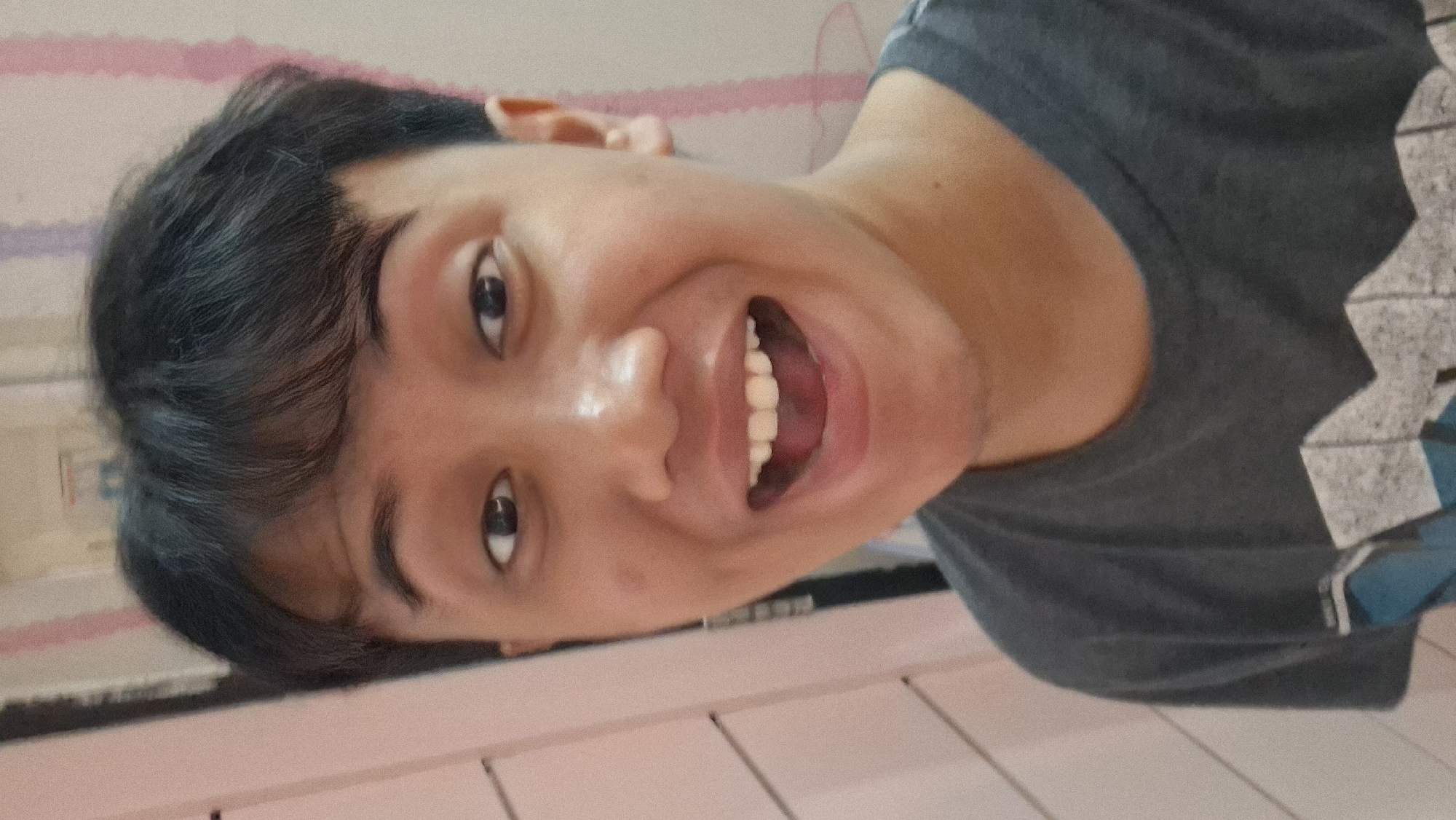 The image displays a close-up shot of a young person, likely male, with dark hair and dark eyes, oriented at a significant tilt. He is looking directly at the camera with a wide, open-mouthed smile, revealing his white teeth and tongue, conveying an expression of joy, excitement, or playfulness.
The subject has light to medium skin, with a slight sheen visible on the forehead and cheeks. He is wearing a dark grey t-shirt with a white and light blue graphic design partially visible on the right side of the chest. His dark hair is styled with bangs across the forehead.
The background is out of focus, featuring a light-colored wall with faint horizontal stripes in shades of pink and light grey/purple at the top, and a lighter, possibly tiled or paneled surface at the bottom. The lighting is bright and appears even, illuminating the subject's face clearly. No specific time of day or outdoor weather is discernible. There is no visible text in the image. The location context provided for this scene is Ciseeng, Indonesia.