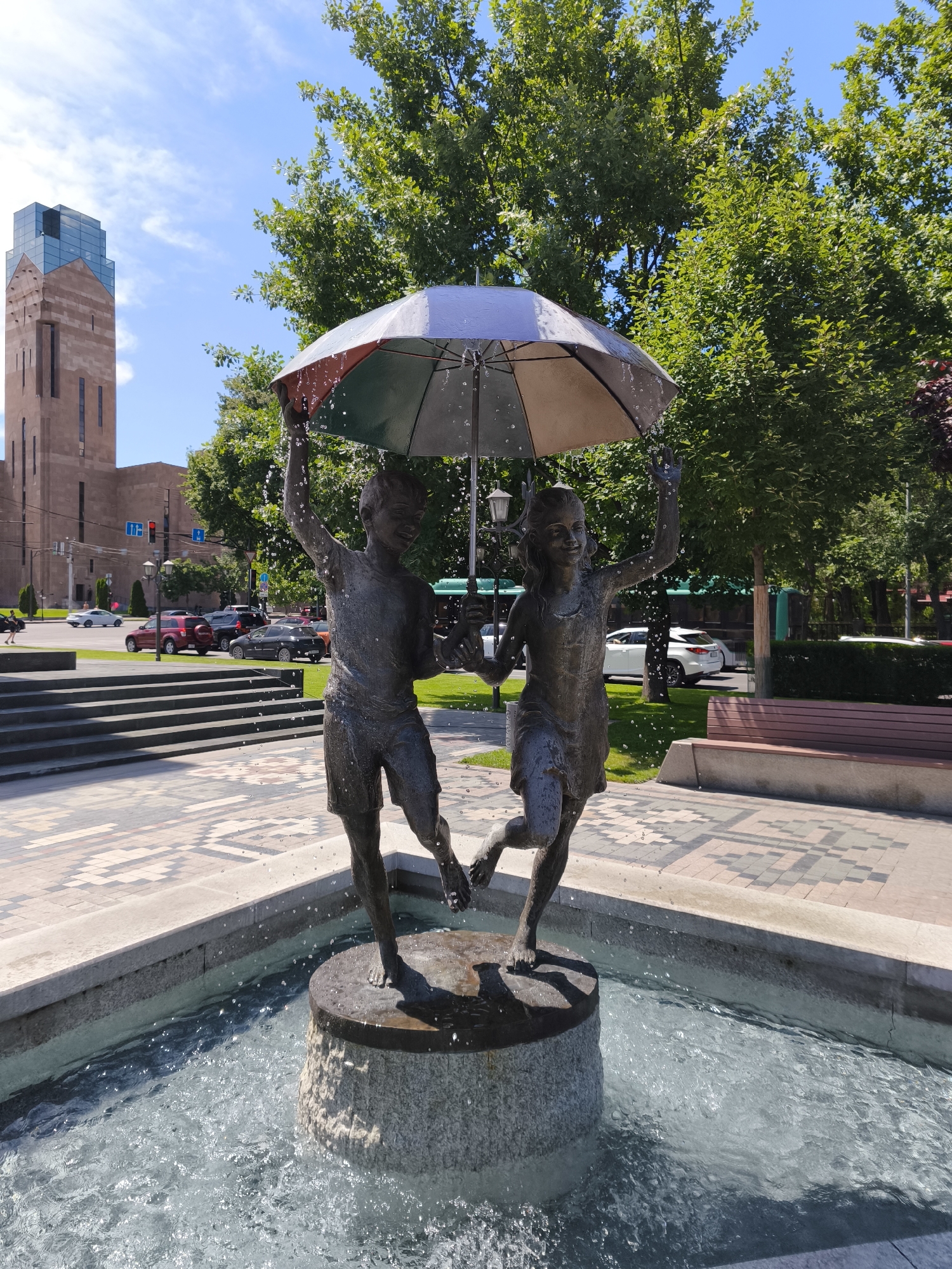 A bronze fountain sculpture depicts a boy and a girl holding an umbrella. Water sprays from the umbrella and around the fountain base. The sculpture is situated in a paved area with patterned tiles, adjacent to a shallow water basin. In the background, a multi-story brick building with a glass facade stands prominently. Trees line the street in front of the building, and several cars are visible on the road. A park bench is located to the right of the fountain. The scene appears to be a public space in Yerevan, Armenia, likely a city square or park.
