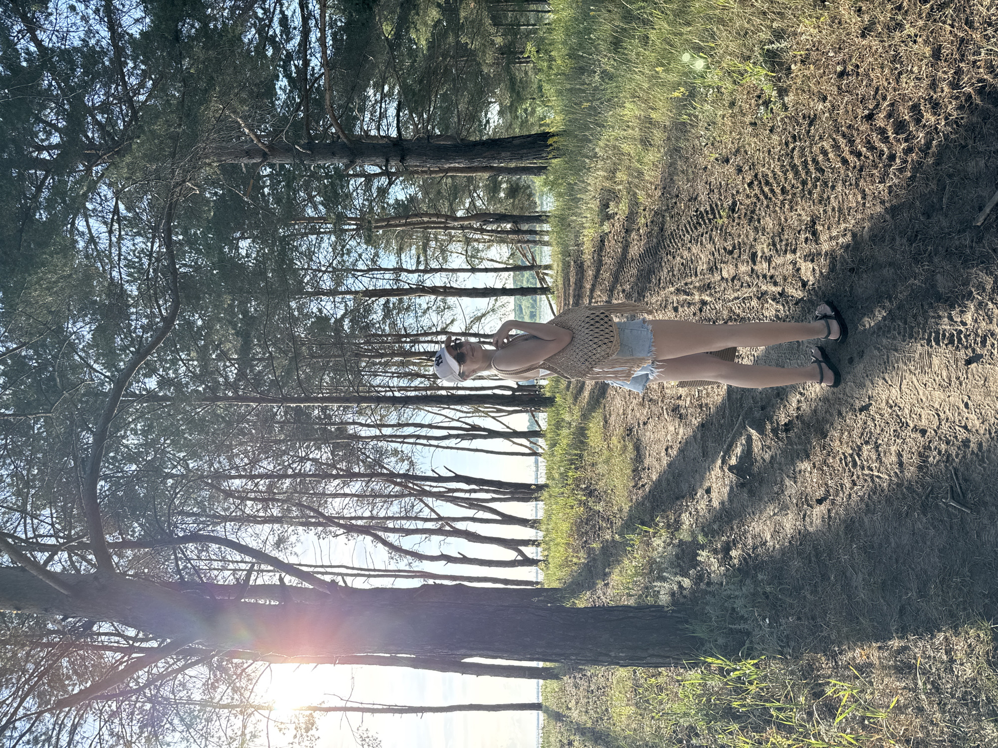 A young woman is standing on a dirt path in a pine forest. She is wearing denim shorts, a white tank top, a white baseball cap, and sunglasses. She has a woven bag with fringe slung over her shoulder. The sun is setting, casting long shadows through the trees. There is a body of water visible in the distance. The path appears to be a hiking trail.