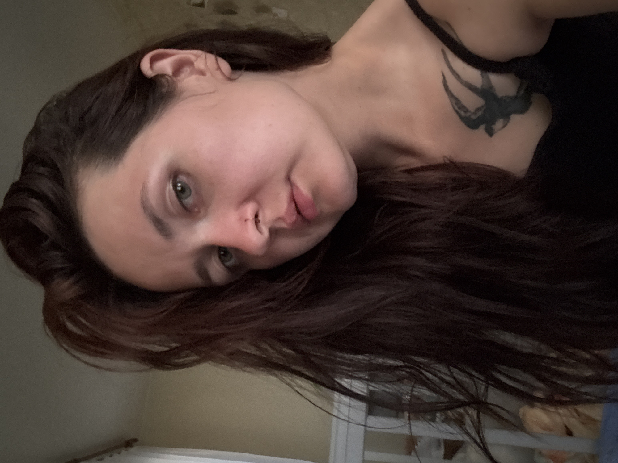 A medium shot shows a woman with long, dark brown hair in a self-portrait indoors. She has green eyes and a small nose ring. She is wearing a black tank top, and a tattoo of a bird is visible on her chest. The background includes a crib and a wall with a pale yellow wallpaper. The lighting suggests it is daytime.