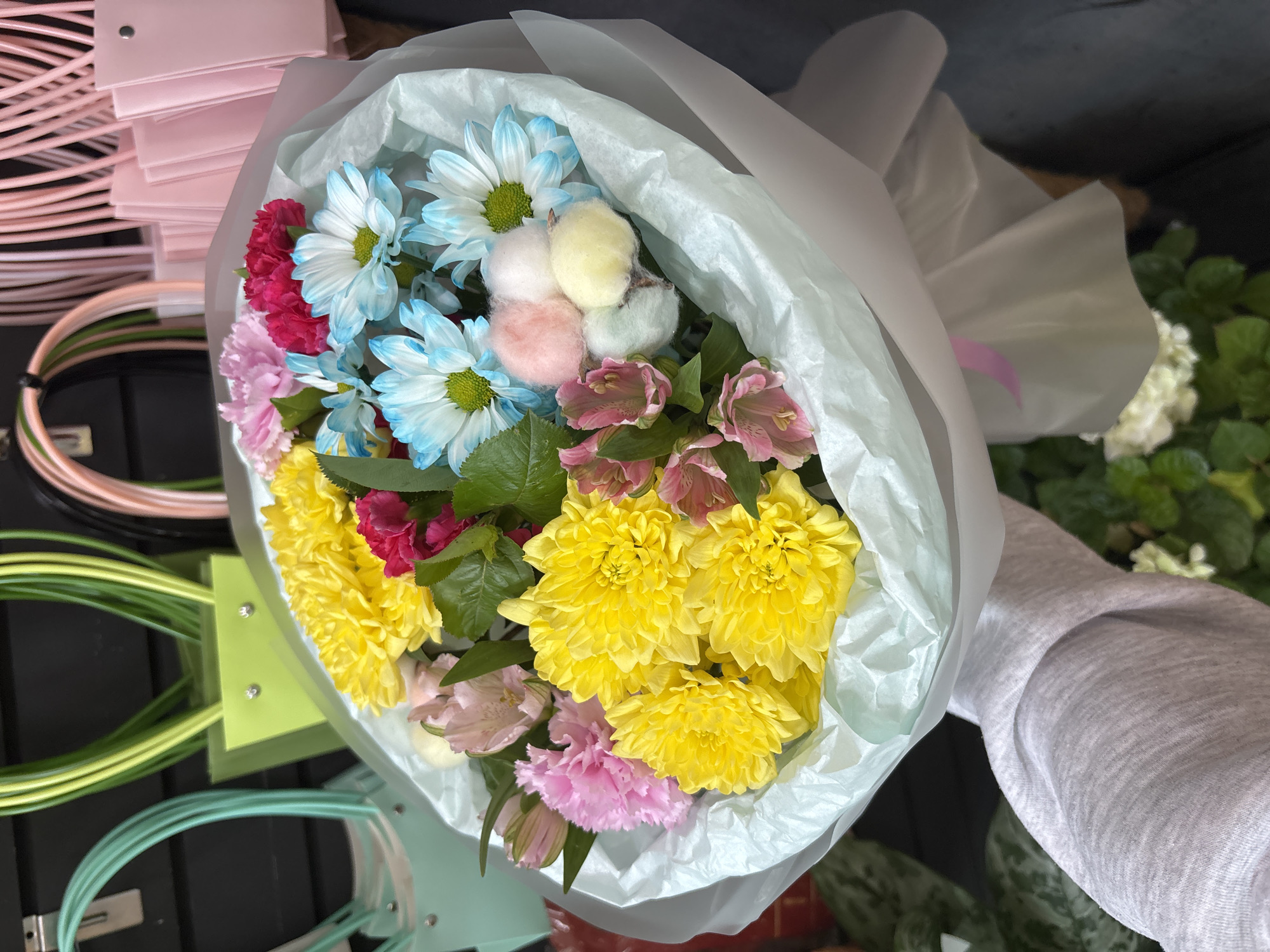 A hand wearing a light gray long-sleeved shirt holds a bouquet of flowers wrapped in light blue and white paper. The bouquet contains bright yellow chrysanthemums, pink carnations, blue and white daisies, and pink alstroemeria. Decorative fluffy pom-poms in pink, white, and yellow are interspersed among the flowers. In the blurred background, several colorful shopping bags with looped handles are visible, including green, pink, and teal. The scene appears to be indoors, possibly a store or market. The image is taken from a slightly low angle, looking up at the bouquet.