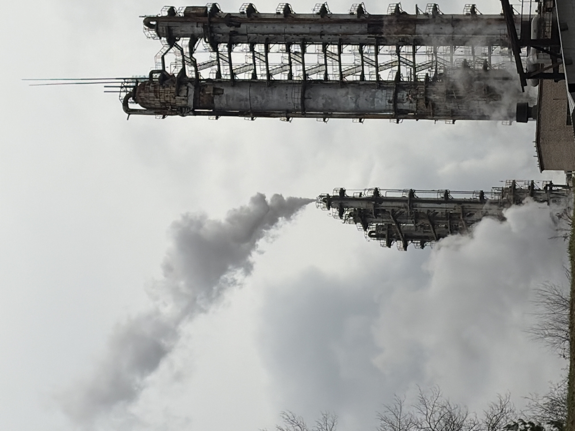 The image shows industrial structures against a cloudy sky. Two elongated, gray metal structures with external scaffolding and ladder systems dominate the upper portion of the image, with the larger structure at the top. Smoke or steam plumes are visible emanating from the structures. Bare trees and vegetation are in the foreground, near the bottom of the image. The scene appears to be an industrial area, potentially located in or near a city, which has been identified as Chernushka, Russia.