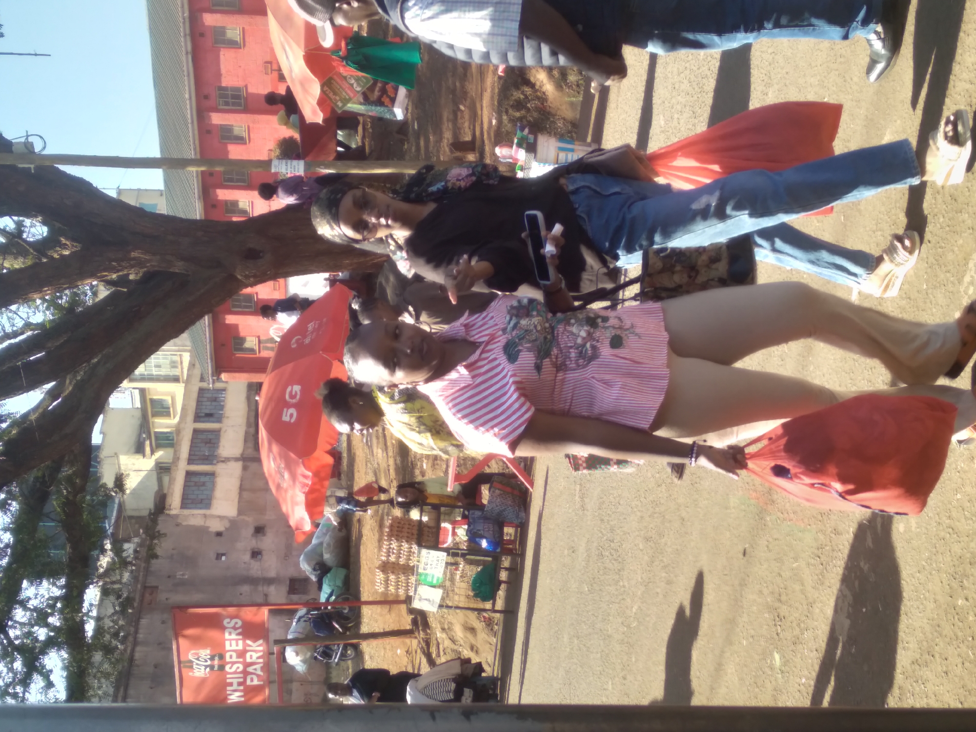 The image depicts an outdoor scene in Nyeri, Kenya, likely a market or roadside stall area, with several individuals and objects visible. In the foreground, two women are walking. The woman on the left is wearing a pink and white striped shirt and beige pants, carrying a red bag. The woman on the right is wearing a dark top, blue jeans, and sunglasses, also carrying a red bag and a purse over her shoulder. She appears to be gesturing with her right hand while holding a mobile phone in her left. Behind them, other people and various items are present, including stalls with merchandise like eggs, umbrellas, and bags of goods. The background features buildings, a large tree, and a sign that reads "WHISPERS PARK". The overall environment suggests a bustling daytime activity in a Kenyan urban setting.