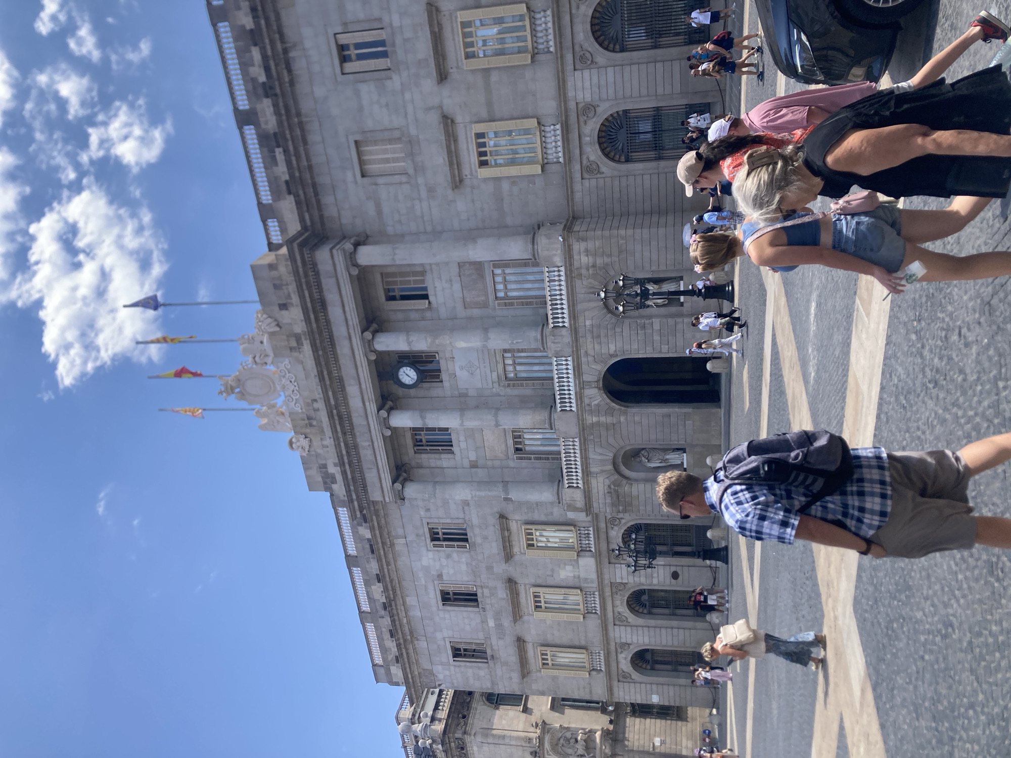 The image depicts the Plaça de Sant Jaume in Barcelona, Spain, a historic square that hosts the Palau de la Generalitat (seat of the Catalan government) and the Ajuntament de Barcelona (City Hall). In the foreground, a man wearing a blue and white plaid shirt, khaki shorts, and a backpack walks away from the camera. To his left, a woman with blonde hair tied back, wearing a beige tank top and blue jeans, walks with a backpack. Behind them, a group of people are also walking across the square. The background features the grand stone facade of one of the buildings surrounding the square, characterized by its columns, arched windows, and a large clock. Several flags are visible on top of the building, including the flag of Catalonia. The sky is a clear blue with scattered white clouds, and the ground is paved with a distinctive checkerboard pattern. The overall scene captures a lively day in a significant public space in Barcelona.
