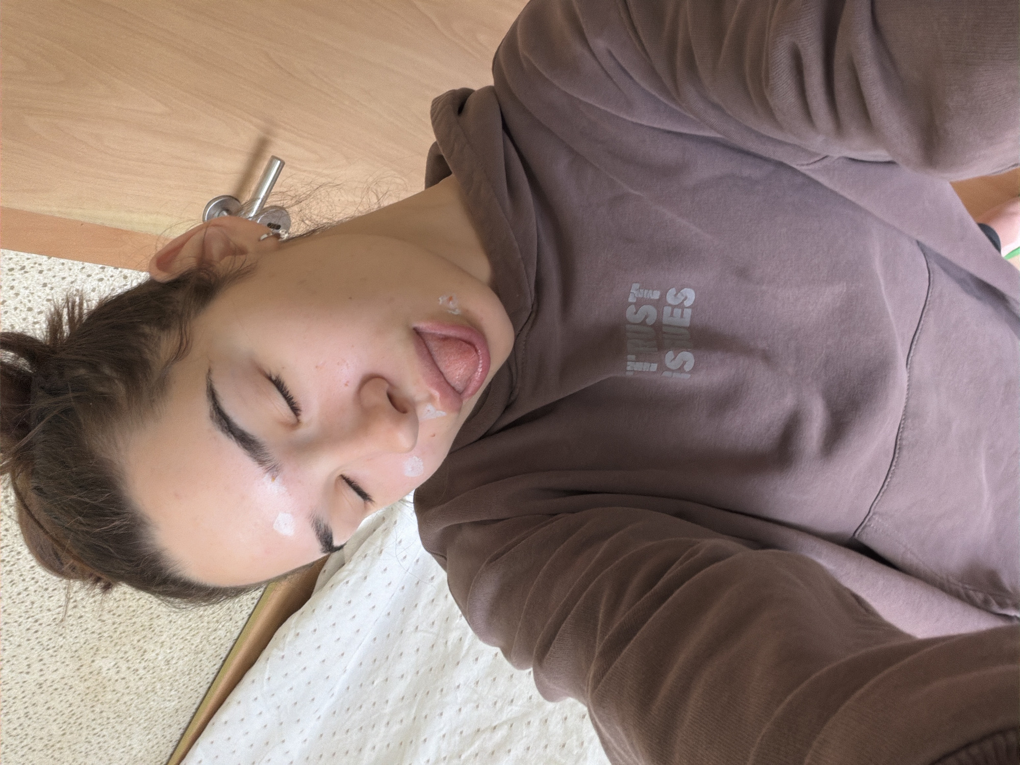 A person with dark hair, tied in a bun, is seen in a close-up, angled selfie, lying down. She is wearing a brown or taupe hooded sweatshirt with the text "TRUST ISSUES" printed in white capital letters across the chest. Her eyes are closed or squinted, and her tongue is slightly out. White, creamy spots are visible on her forehead, cheek, and around her nose and mouth, suggesting a skincare product or playful application of a substance. The background features a light wood surface, a metallic door handle, and a white textured fabric, likely a blanket or sheet, where she rests. The indoor lighting is soft. The scene evokes a casual, intimate moment, possibly during relaxation or a beauty routine. No specific location cues for Sofia, Bulgaria are visible in the image.