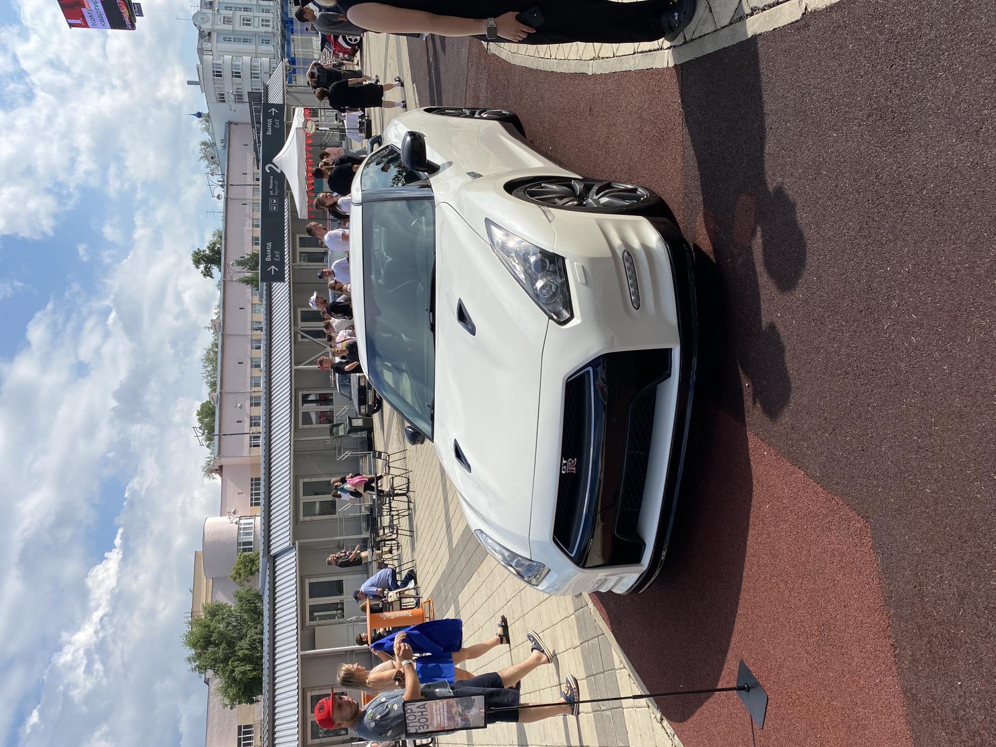 A white Nissan GT-R is parked on a red rubberized surface, positioned diagonally. The car's front is angled towards the viewer, showcasing its headlights, grille with the GT-R emblem, and black lower bumper. In the background, a white building with a metal roof runs horizontally across the image. Several people are visible in front of this building, some standing, others walking. One man in a red cap and shorts stands to the left of the car, looking towards it, holding a blue cloth. A woman in a light-colored top and a blue skirt walks past him. Further back, a white tent with red trim is set up. The sky above is partly cloudy. The scene appears to be an outdoor event, possibly a car show, taking place in a city. Based on contextual clues, the location is the exhibition center "Expobel" in Yekaterinburg, Russia.