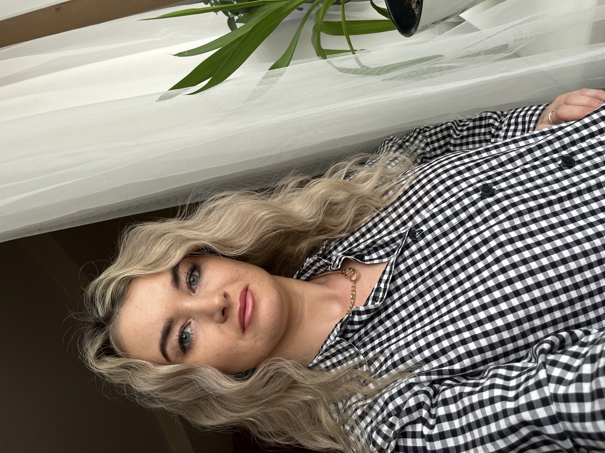 A woman with long blonde curly hair is wearing a black and white checkered button-down shirt and a gold chain necklace. She is looking at the camera with a neutral expression. She is indoors with a white sheer curtain and a plant in the background. The exact location in Chaikovsky, Russia cannot be determined from the image.
