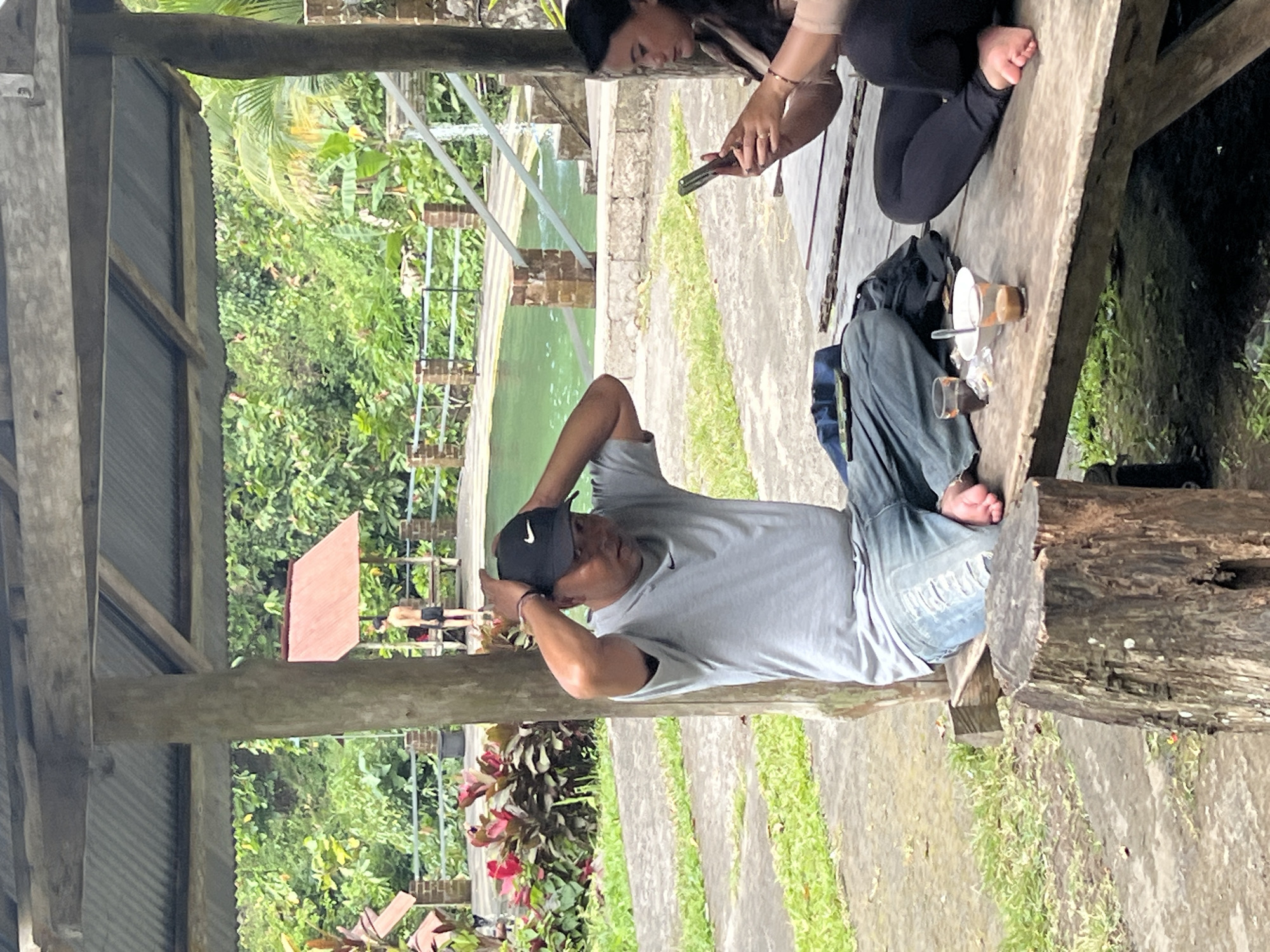 The image depicts an outdoor scene under a wooden shelter with a corrugated metal roof. In the foreground, a man wearing a grey t-shirt and blue jeans is seated on a wooden bench, with his hands on his head, wearing a black baseball cap. To his right, a woman in black pants and a light-colored jacket is also seated on the bench, looking at her phone. On the bench between them are two glasses of beverage and a white bowl. In the background, there is a swimming pool with green water, surrounded by lush vegetation and a brick and metal railing. A young man in black shorts is standing near a red-roofed structure in the distance. The ground is paved with concrete slabs interspersed with grass. The overall setting suggests a recreational area, possibly a resort or park in Sukasada, Indonesia.