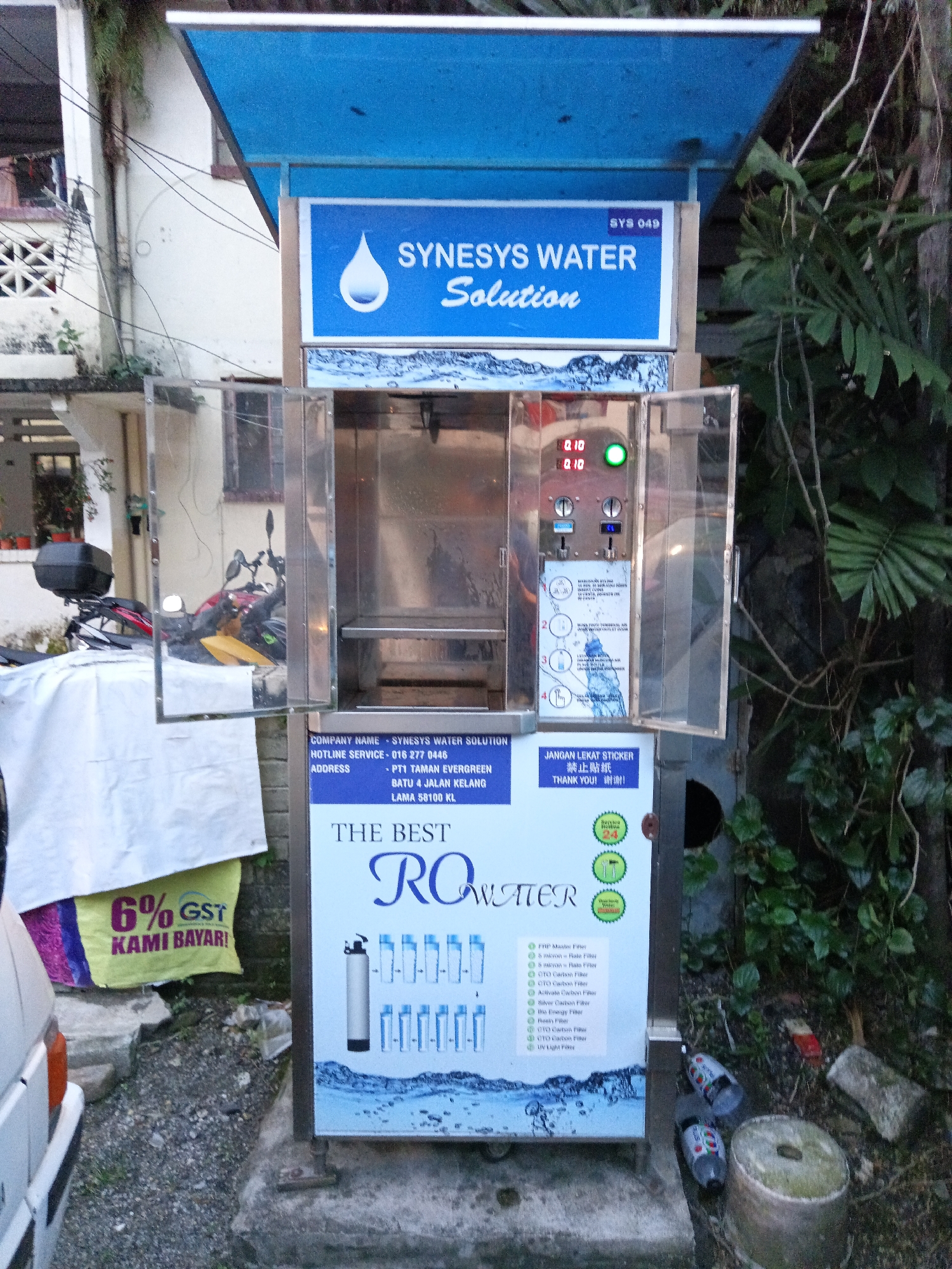The image shows an outdoor water dispenser machine, likely located in Kuala Lumpur, Malaysia, given the address text which includes "BATU 4 JALAN KELANG LAMA 58100 KL". The machine has a blue canopy and a prominent blue sign with "SYNESYS WATER SOLUTION" written in white text. Below this, there is a compartment with glass doors revealing the internal mechanism of the dispenser, including two digital displays showing "0.10" and a green indicator light.
To the left of the dispenser, there is a white sheet covering an object, possibly a motorcycle, with a black box on the back. Next to this sheet, a yellow banner with "6% GST KAMI BAYAR!" is visible.
The lower part of the machine features a large graphic advertisement for "THE BEST RO WATER", listing various types of filters. To the right of the dispenser, there is a dense green foliage and some discarded plastic bottles on the ground. The ground surface appears to be a mix of gravel and concrete. The overall environment suggests a somewhat unkempt urban outdoor setting.