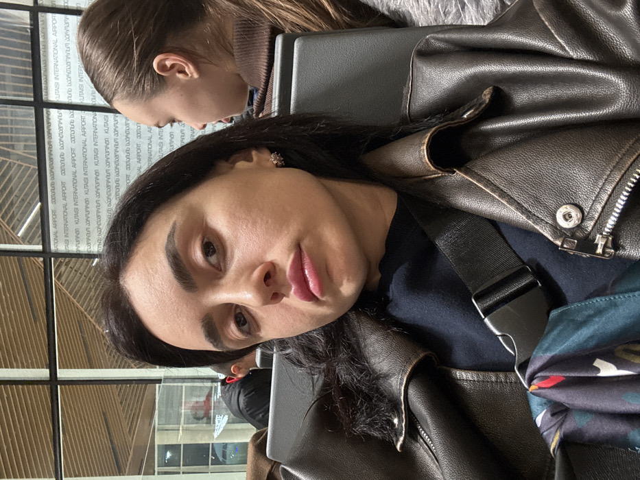 A woman with dark hair and a leather jacket poses for a selfie at what appears to be an airport, as evidenced by the writing in the background that contains the word "airport." Another person with brown hair sits behind her. The woman in the foreground wears a black strap across her chest. The environment around them appears to be well-lit and enclosed.