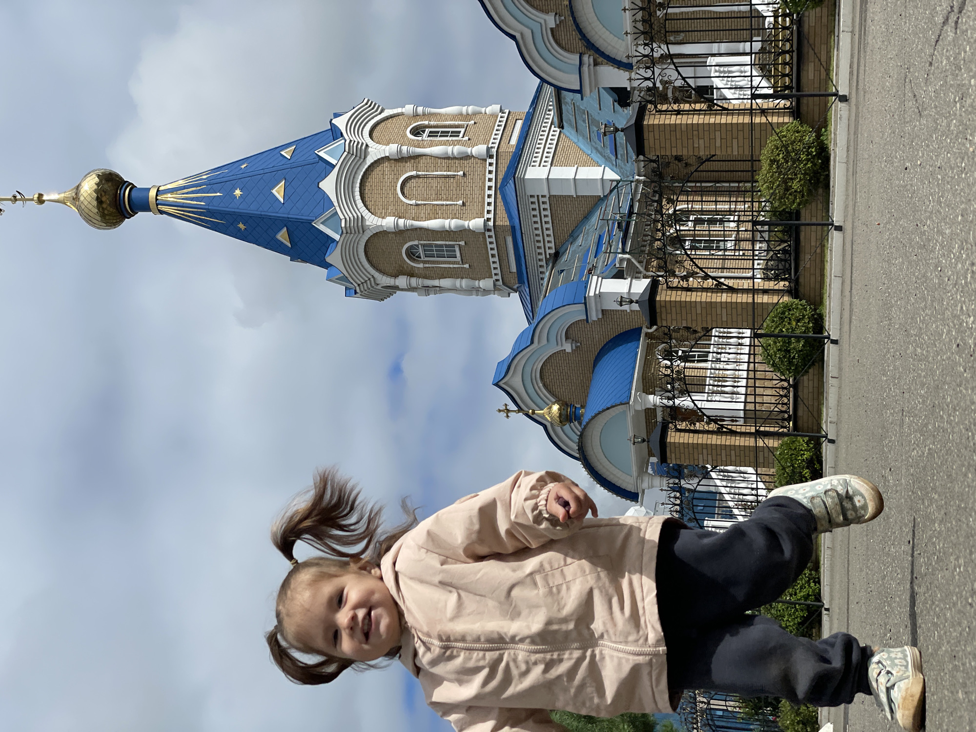 In Izhevsk, Russia, a young girl with two pigtails is captured mid-stride, her face lit with a joyful expression. She is wearing a light pink hooded jacket and dark pants, with floral-patterned sneakers. The background features the distinctive blue and gold domes of the St. Michael's Cathedral. The cathedral, a prominent landmark in Izhevsk, is partially visible, with its ornate architecture including brickwork and decorative white elements. A black wrought-iron fence with brick posts separates the sidewalk from the church grounds, and manicured green bushes are visible behind the fence. The sky above is cloudy, suggesting overcast weather.