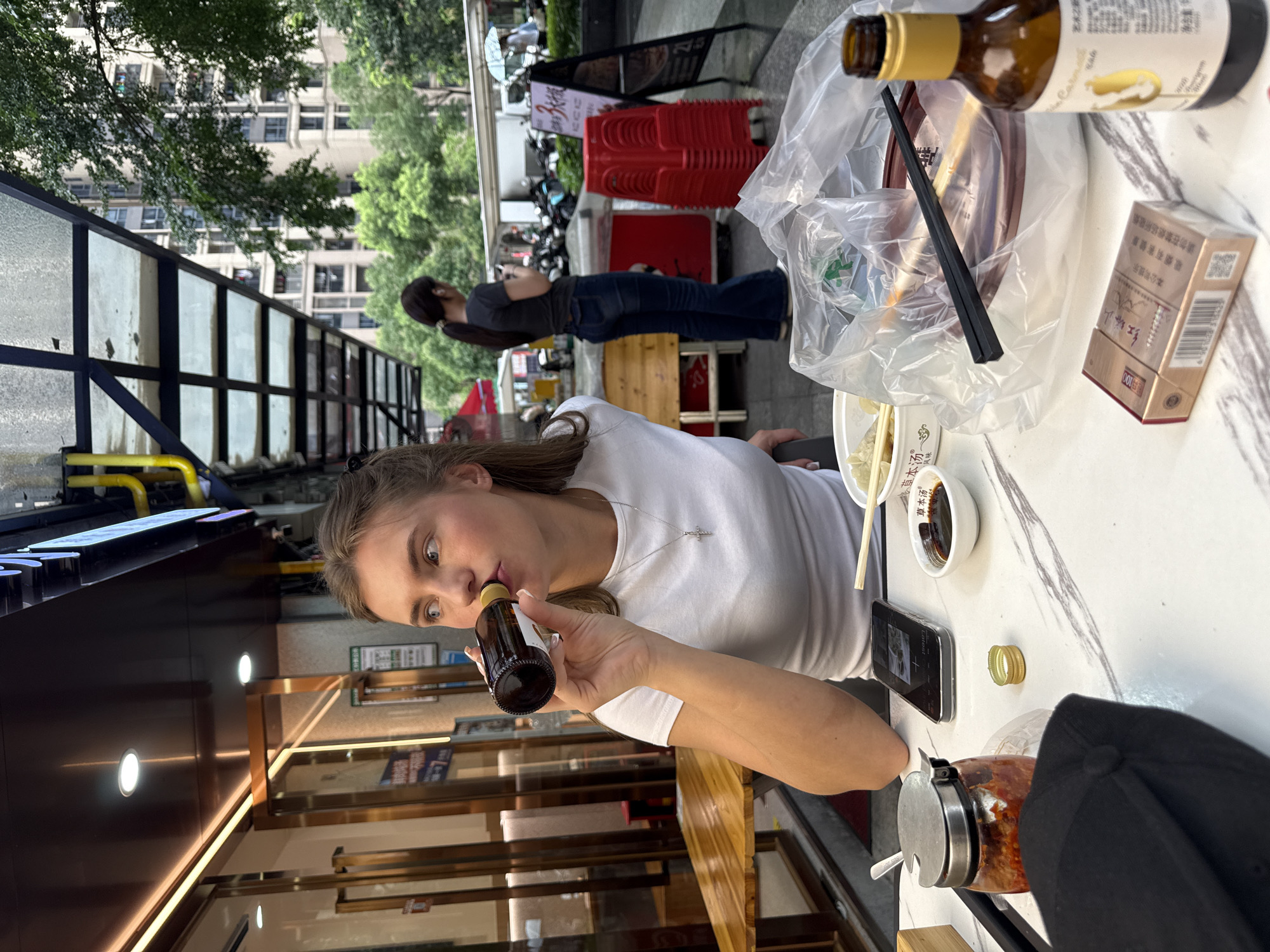 A young Caucasian woman with blonde hair pulled back is seated at an outdoor restaurant table in Haizhu District, China. She is wearing a white t-shirt and a silver cross necklace and is drinking from a dark glass bottle. On the marble-patterned table in front of her are various items including a black baseball cap, a jar of red chili paste, a smartphone displaying an image, a small bowl of dark sauce, a pack of cigarettes, a bottle of amber liquid with a yellow label, a plastic bag containing food containers and chopsticks, and a bowl of what appears to be dumplings. In the background, a second woman with dark hair tied in a ponytail is standing, looking at her phone. The restaurant appears to be on a street with a covered walkway and trees. Other people, motorcycles, and red plastic chairs are visible in the background. The weather appears overcast, possibly rainy.