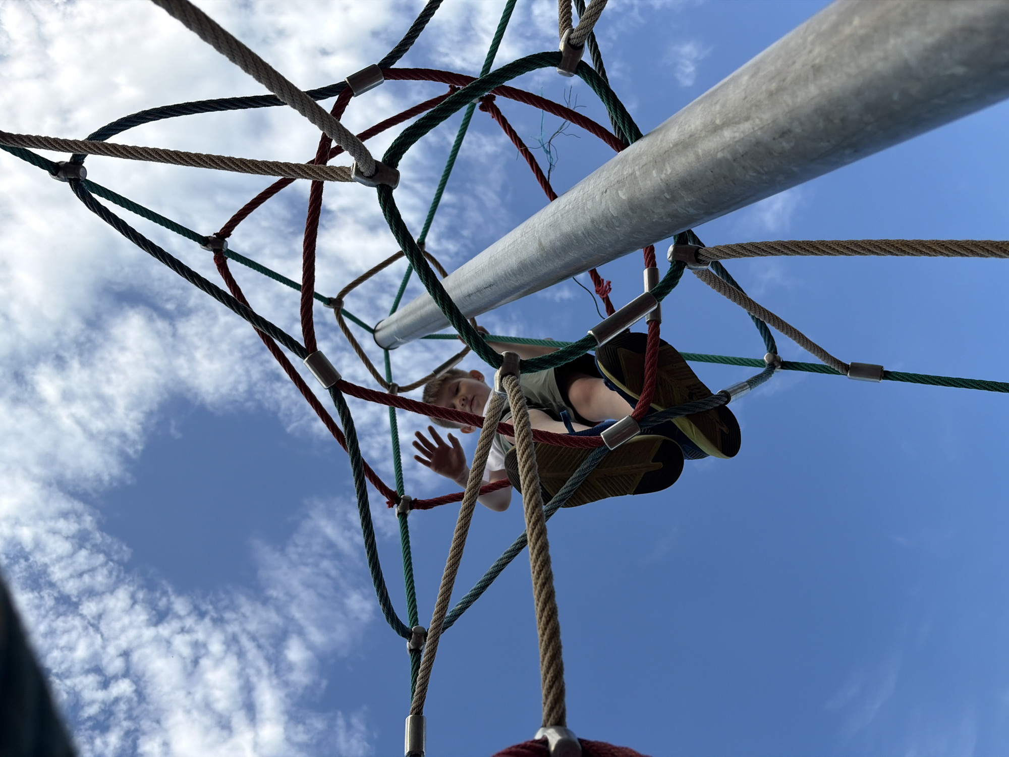 A low-angle shot depicts a child climbing on a rope climbing structure against a bright blue sky with wispy white clouds. The child is positioned within the structure, with parts of their body and shoes visible through the ropes. The climbing structure is made of a network of intertwined ropes of different colors, including red, green, and brown, connected by metal clamps. A large silver metal pole runs through the center of the frame, close to the camera's perspective. The image is taken in an outdoor setting, likely a playground or recreational area, in the city of Unknown, Unknown.
