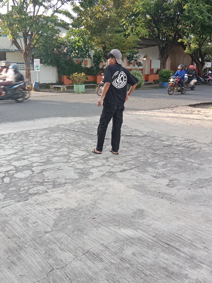 The image shows a young man wearing a black t-shirt with a white logo and black pants standing on a cracked, gray concrete surface. He is looking to the left and has a gray baseball cap on his head. The man is located in Tugu, Indonesia, at the coordinates -6.973 latitude and 110.326 longitude. There are two motorcycles parked in the background, one on the left and one on the right, and another motorcycle is driving away from the man, to the left. There are green bushes and trees in the background. The concrete is in a worn and damaged condition. The man is standing with his right leg slightly bent, as if he is about to take a step forward. The scene is outdoors and the sun is shining. The image is taken at a low angle, making the man appear larger than he is in reality.