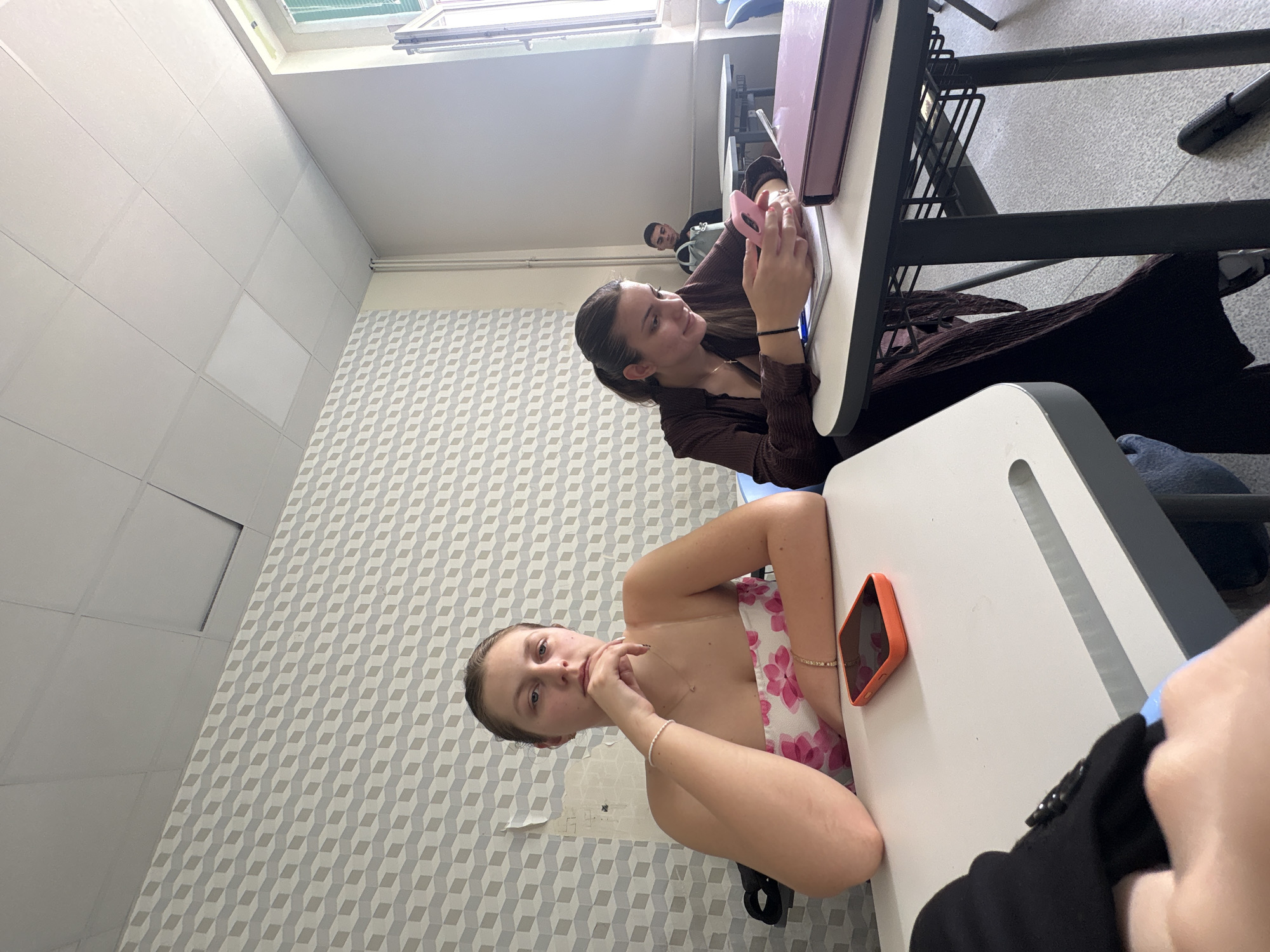 The image displays an indoor scene, likely a classroom or study area in Burgas, Bulgaria, captured from an angled perspective. Two young women are prominently featured. In the foreground, a young woman with light brown hair tied back is seated at a white desk, wearing a white strapless top with a pink floral pattern. She looks towards the camera with a neutral expression, her right hand resting near her chin. An orange-cased smartphone is visible on the desk beside her.
In the middle ground, another young woman with dark hair, dressed in a brown striped top, is seated at a similar desk. She is engrossed in a smartphone with a pink case, which she holds with both hands. Further back, a young man is partially visible, also seated at a desk, looking generally in the direction of the foreground subjects.
The room's walls are predominantly white, with a striking section covered in a light grey and white 3D cube-patterned wallpaper. Bright natural light streams in from a window at the top of the frame, indicating daytime. The furniture consists of modern white desks with black metal frames. A heating pipe runs along one of the walls. The overall setting suggests a casual moment within a learning environment, possibly during a break, with individuals engaged in their own activities. No visible text is clearly discernible.